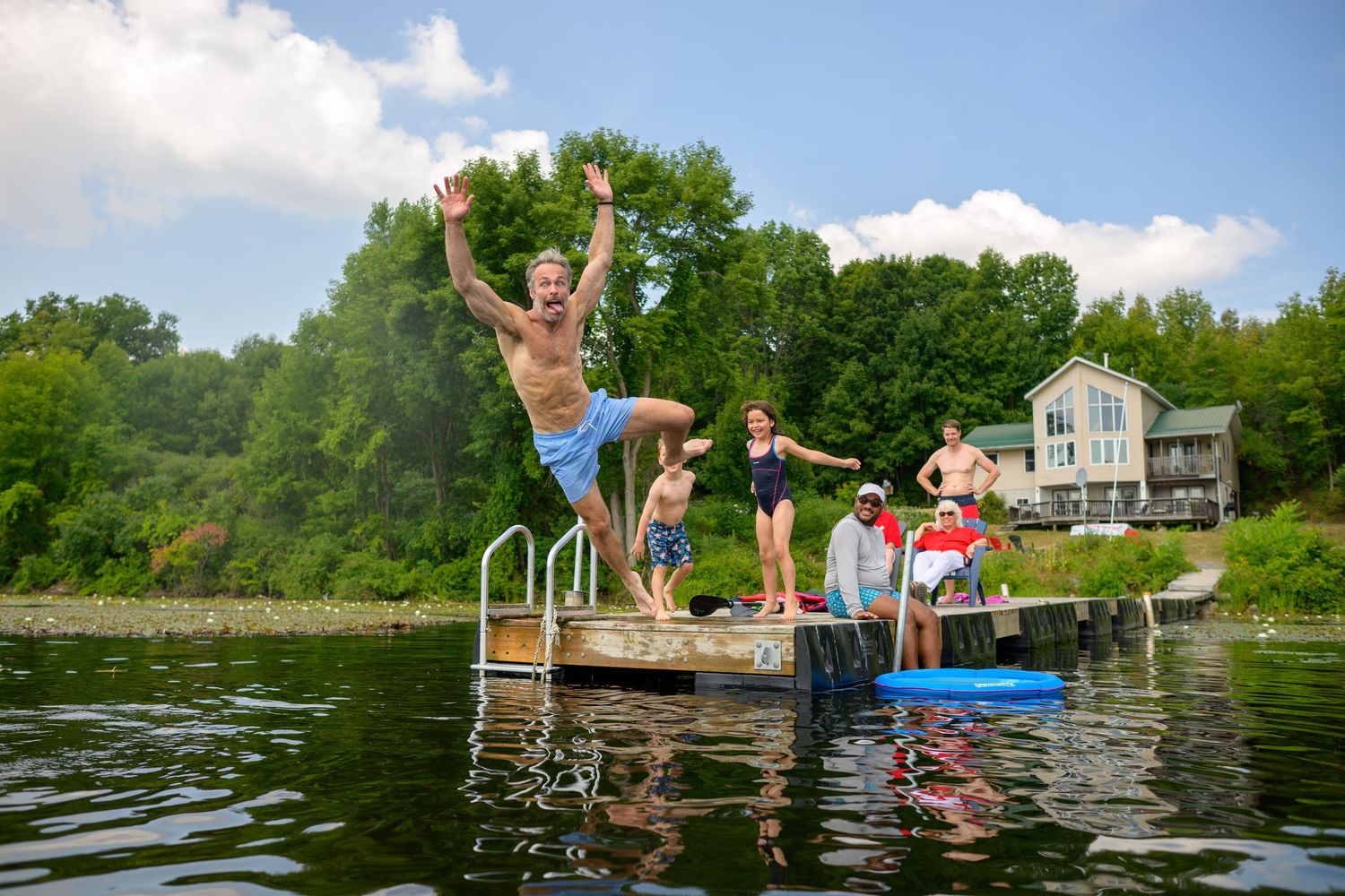 People enjoying summer fun jumping into a lake from a dock with trees and a house in the background.