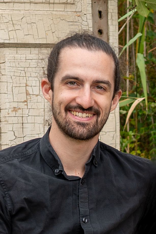 Luke, the photographer, in a black button-up shirt smiling against a light coloured wall.