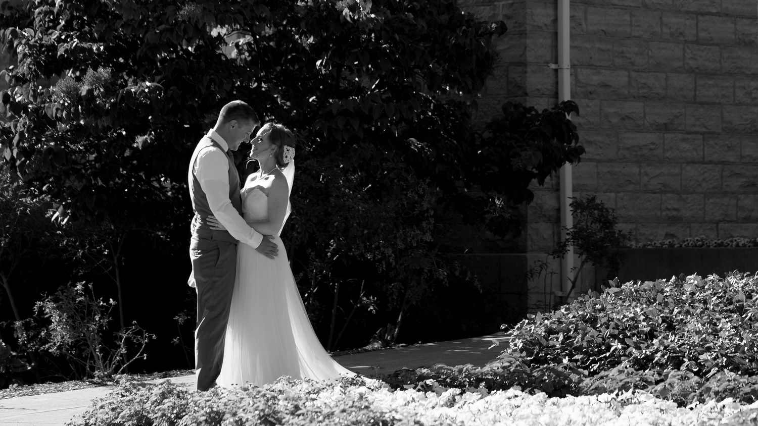 A wedding party poses in front of a historic stone building and fountain on a sunny day.