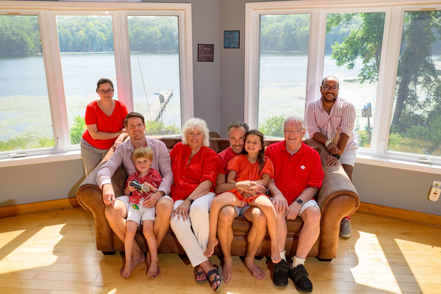 A group wearing matching red shirts sits together on a tan couch in a sunny room with large windows overlooking water.