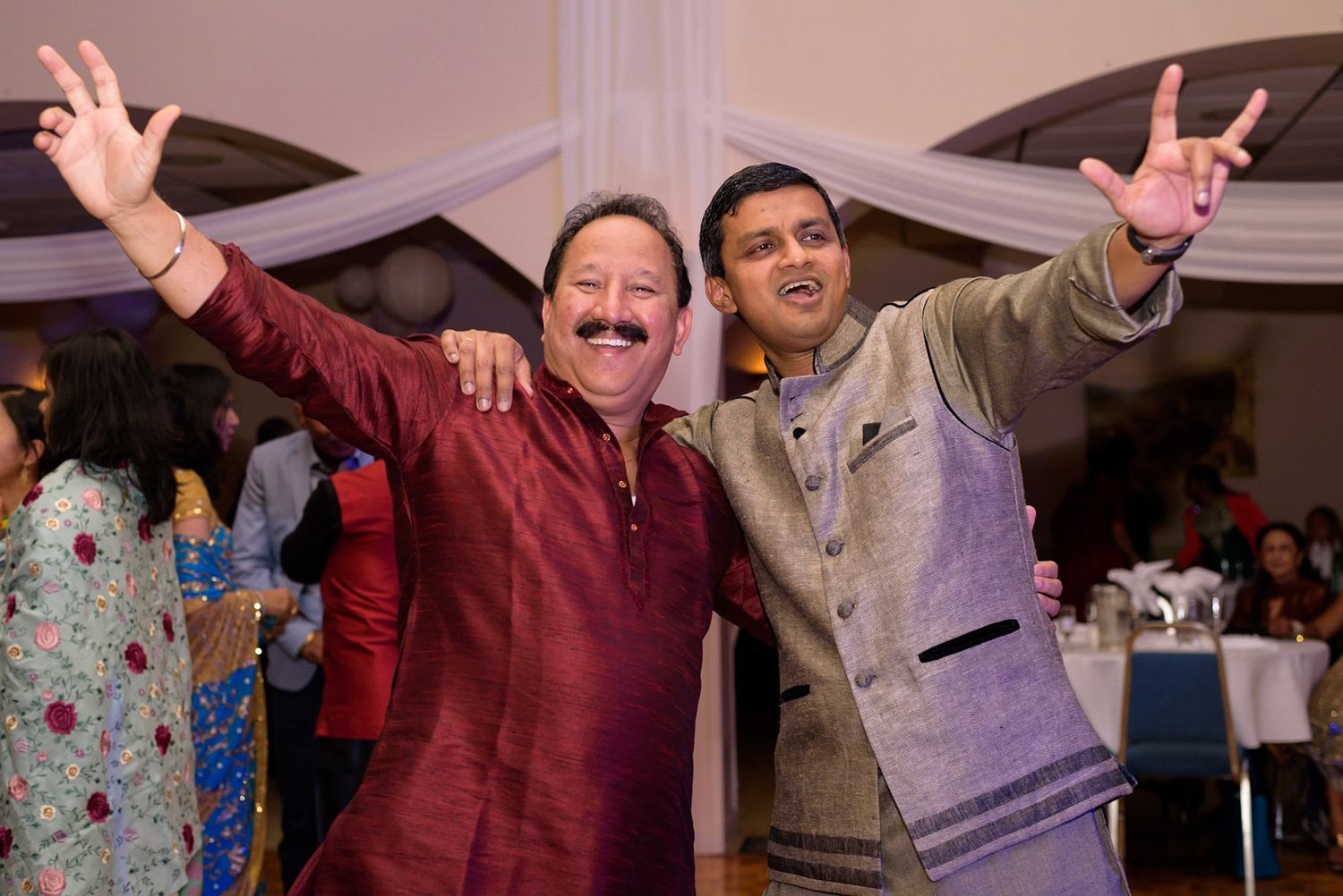 Two men in formal ethnic wear raise their arms in celebration at an indoor event.