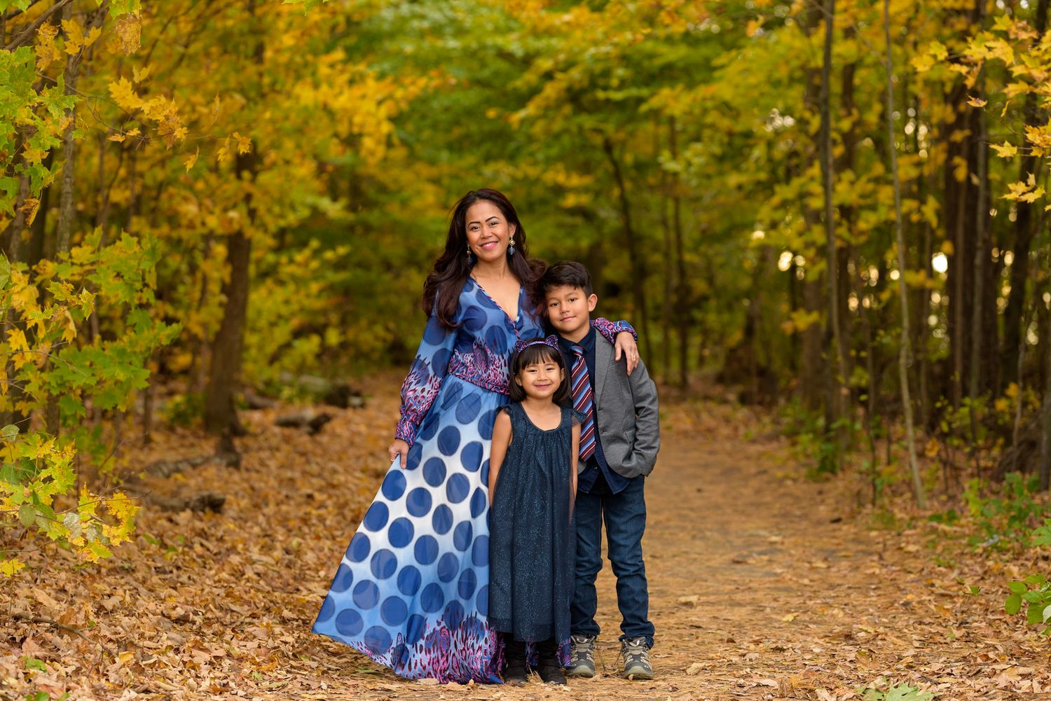Family poses together on a leaf-covered forest trail surrounded by golden autumn foliage.