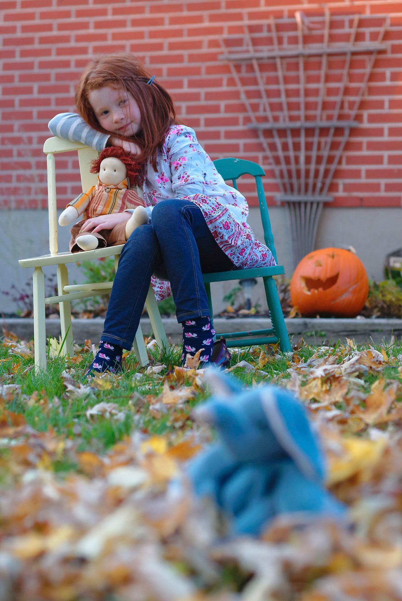 A person sits on a green bench surrounded by autumn leaves and a carved pumpkin decoration in front of a brick wall.
