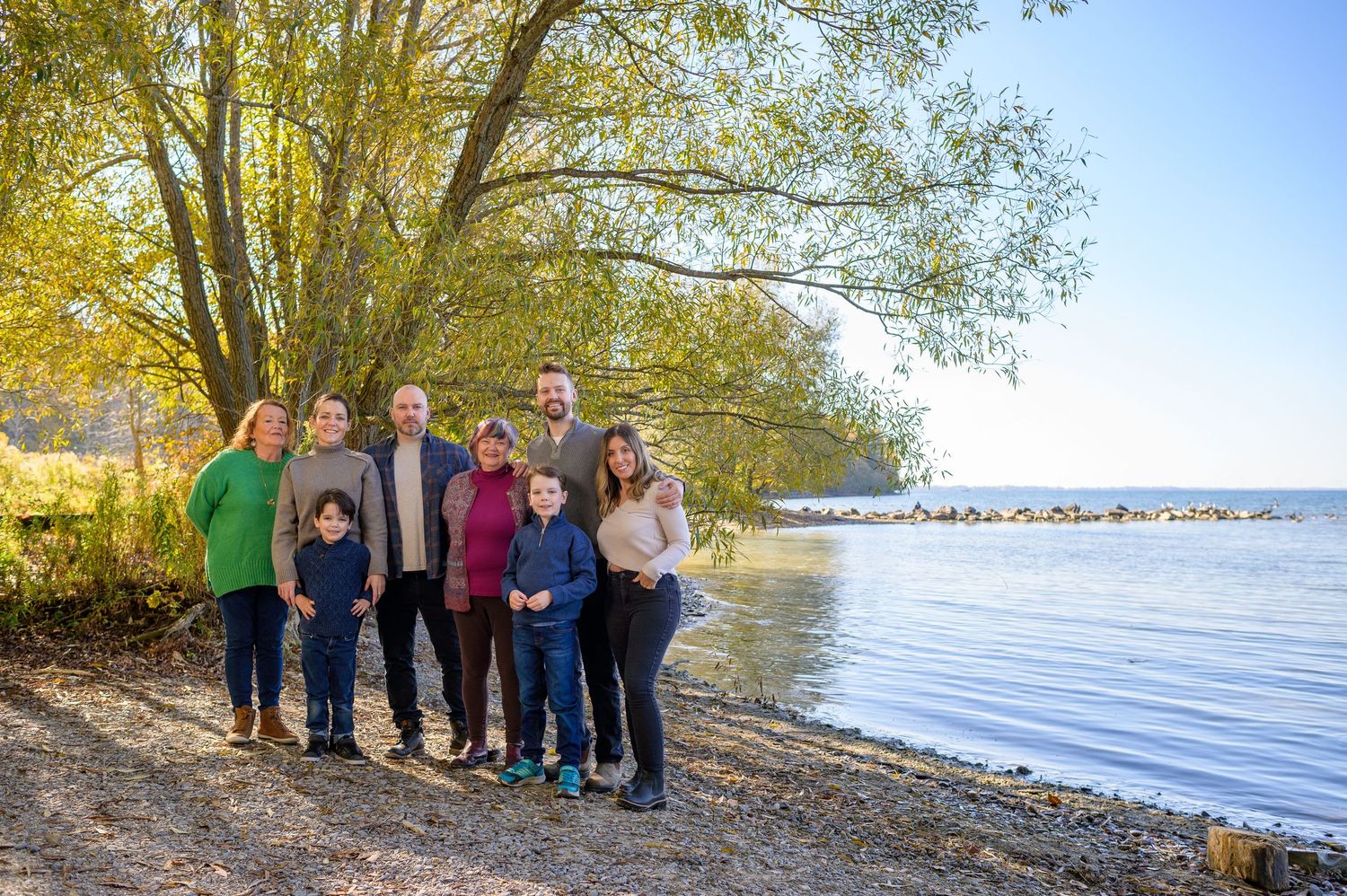 A group of people standing on a rocky beach shore with autumn trees and Lake Ontario in the background.