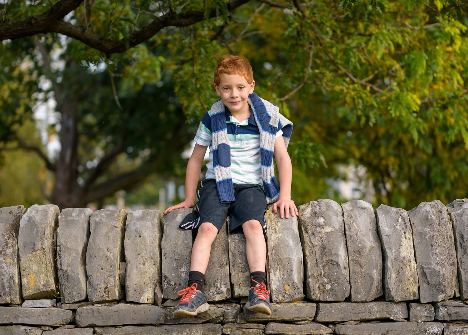 A child sits on a rustic stone wall in a garden setting wearing casual clothing and sneakers.