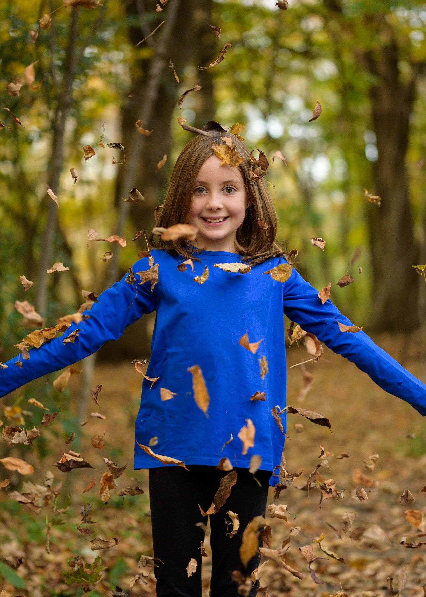 A person in a bright blue sweater plays in falling autumn leaves against a wooded forest backdrop.