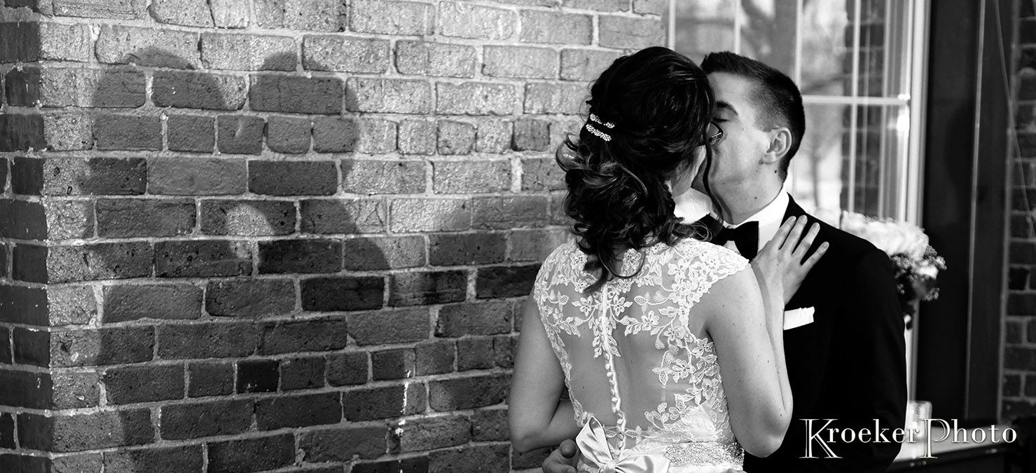 Black and white wedding photo showing couple silhouetted against brick wall with romantic lighting.