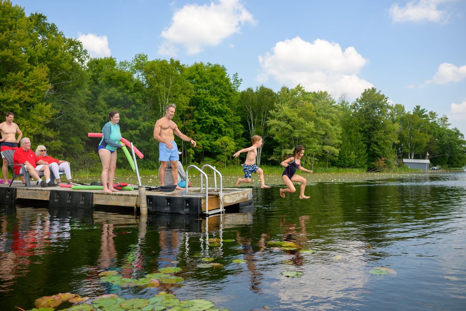 People enjoy jumping into a calm lake from a wooden dock on a sunny summer day.