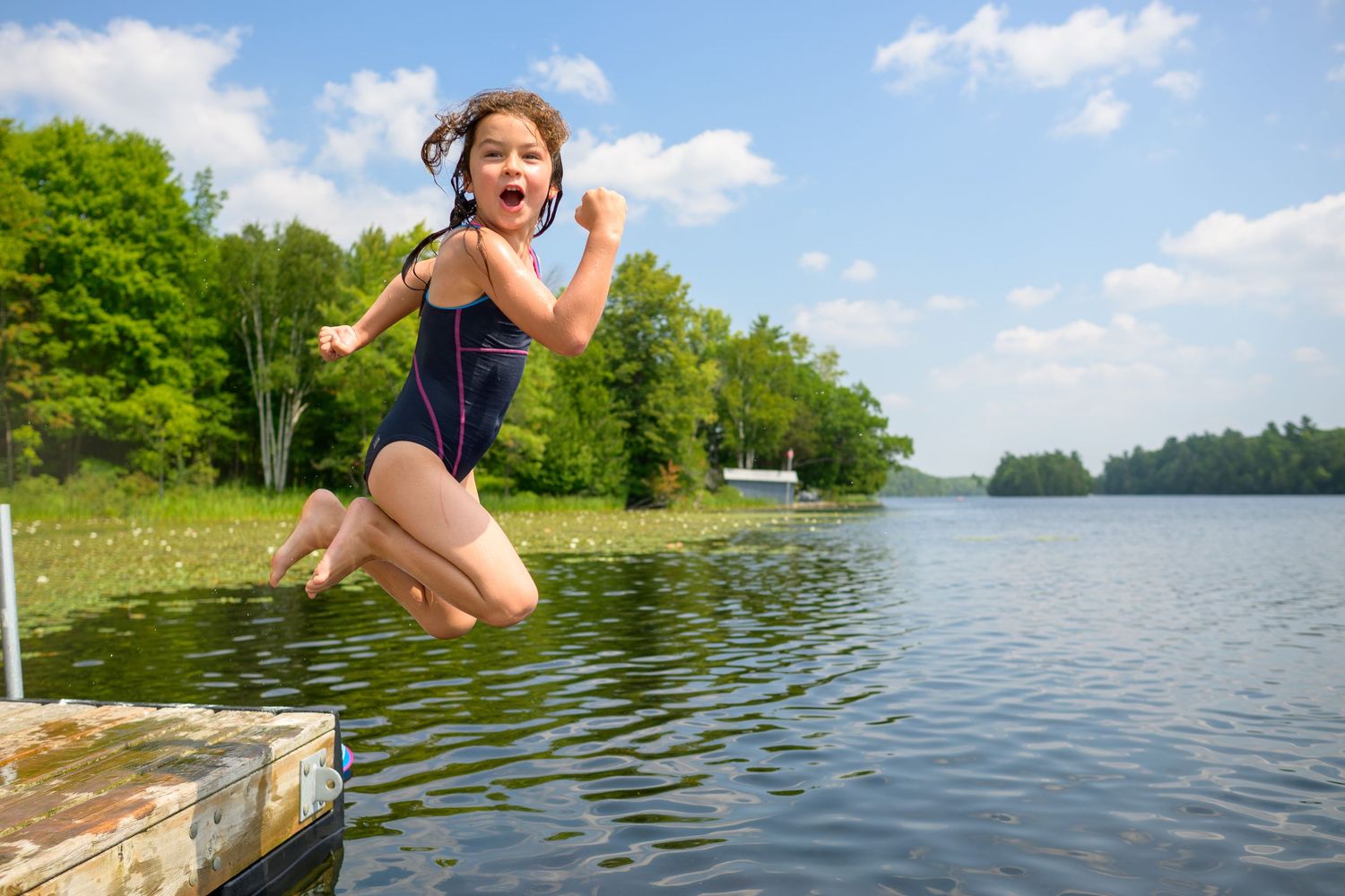 Someone jumps into a scenic lake on a sunny summer day with lush green trees in the background.