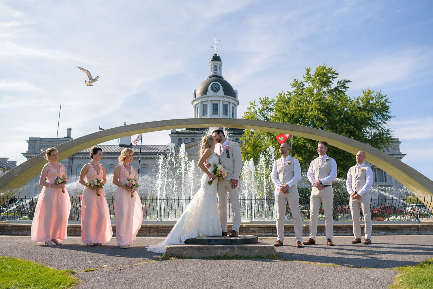 Wedding party members stand in line near an ornate gazebo and fountain.