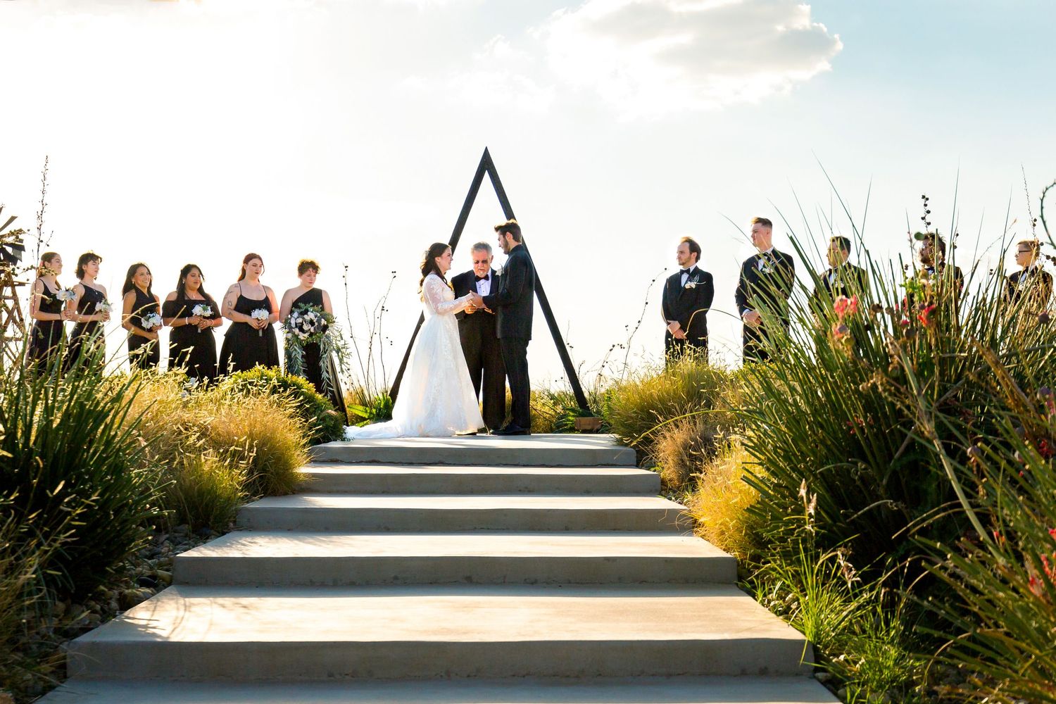 Wedding ceremony taking place on elevated steps with a triangular arch and ornamental grasses on either side.