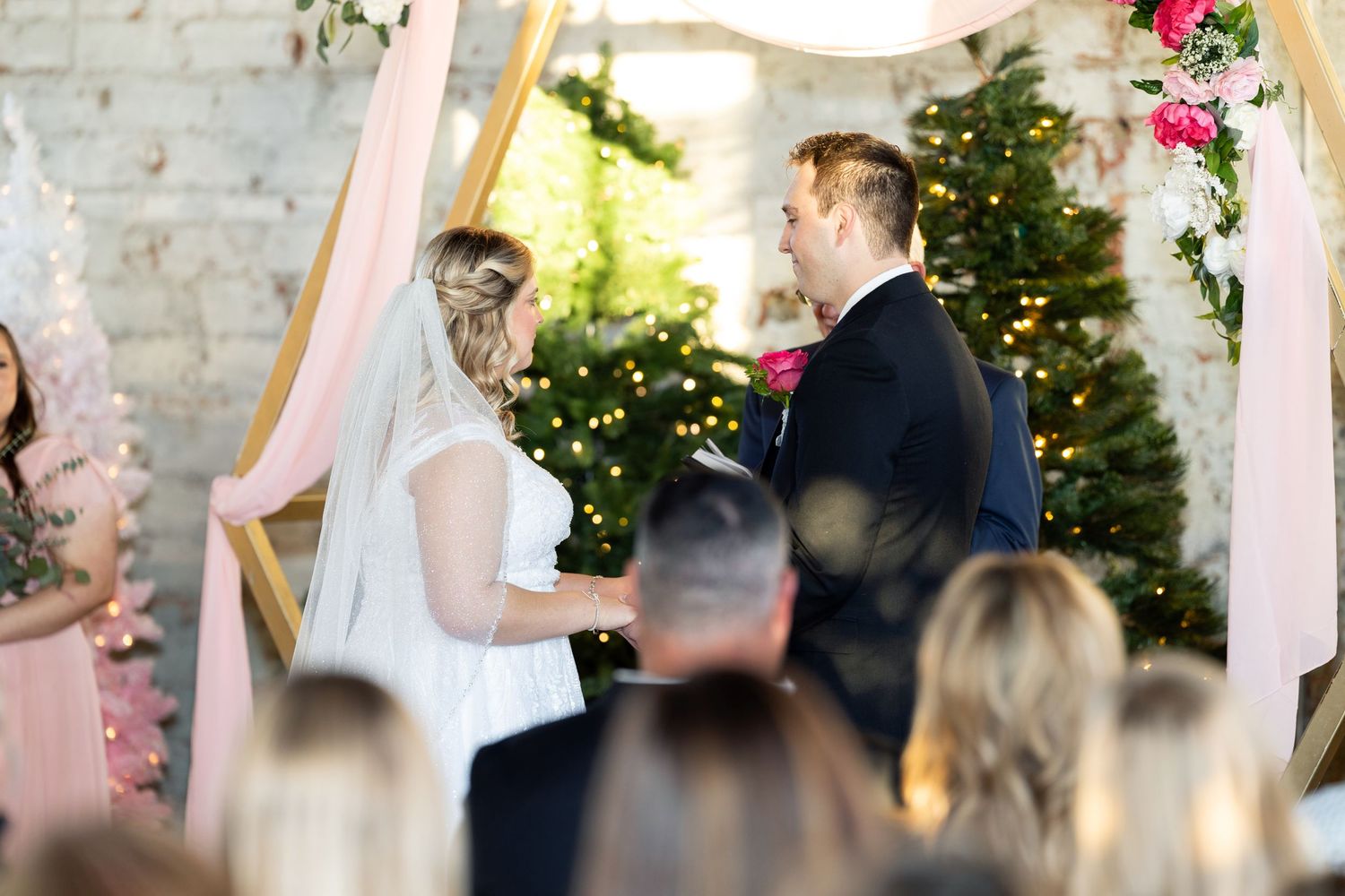 Wedding ceremony under floral arch with pink drapery and Christmas trees in background.