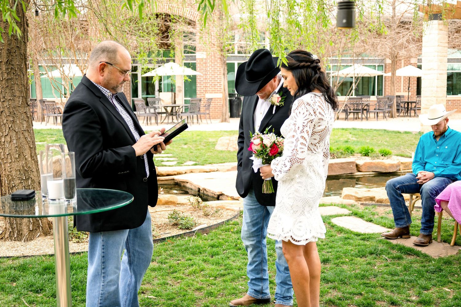 Outdoor wedding ceremony setup with guests gathered on a grassy lawn surrounded by trees.