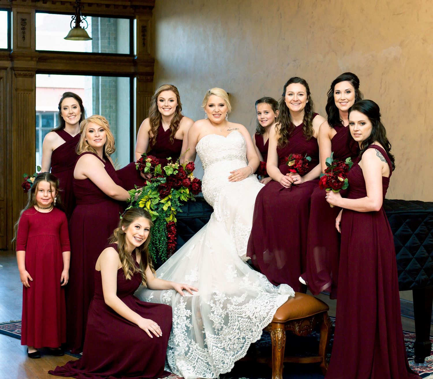Wedding party in burgundy dresses gathered around seated bride in white gown.