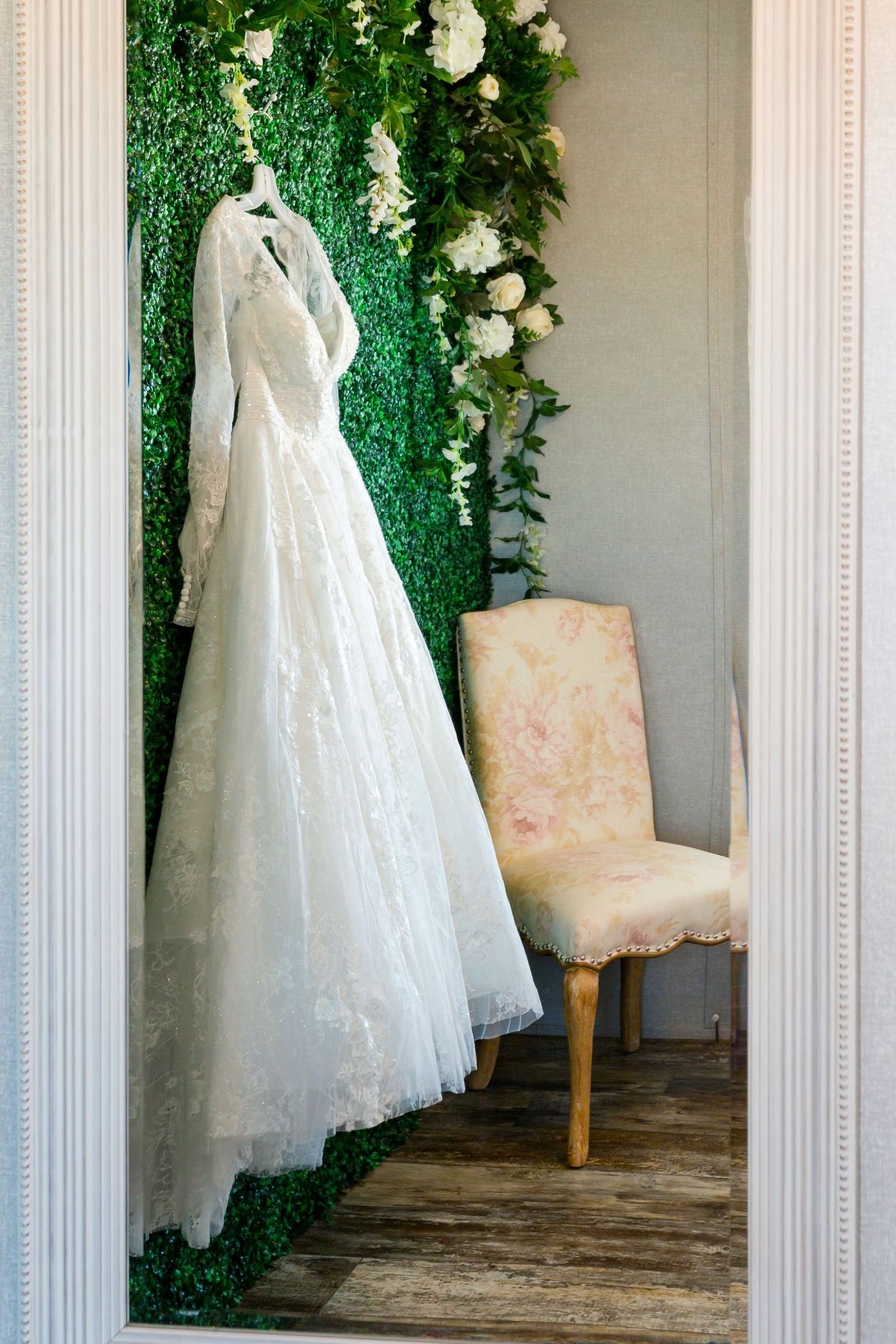 An elegant white lace wedding dress hangs against a green floral backdrop next to an upholstered chair.