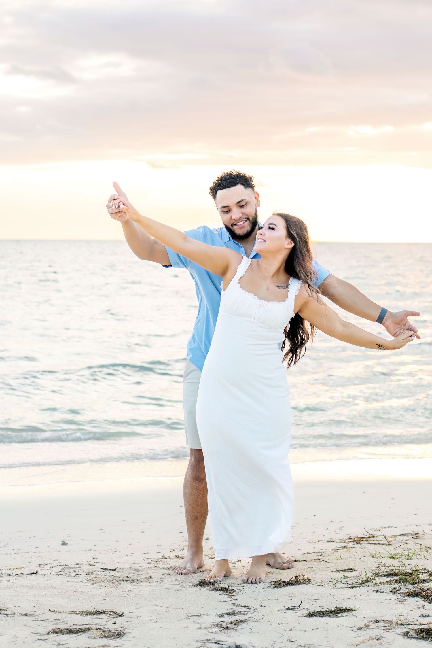 A couple dances playfully on a beach at sunset in white outfits.