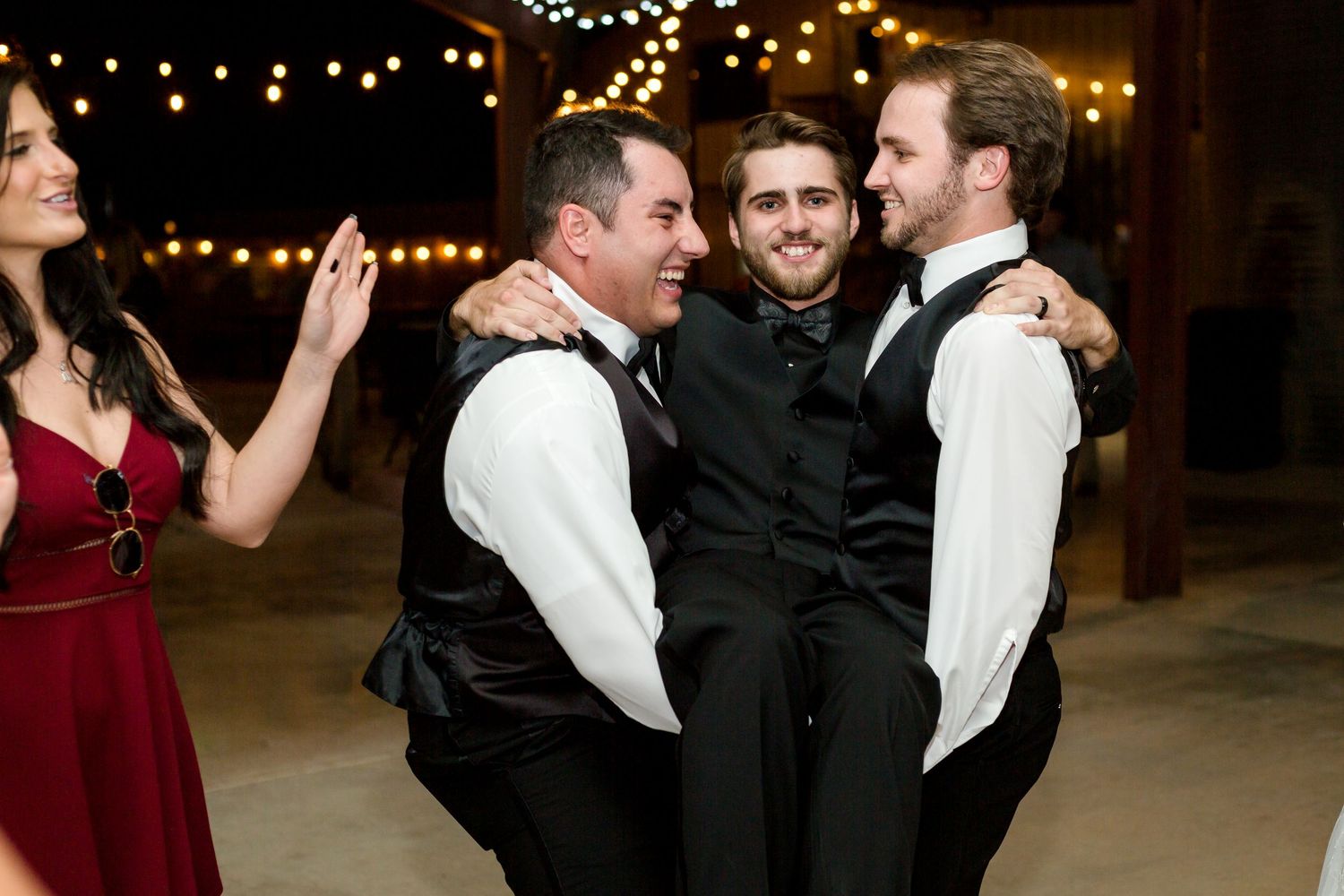 Groomsmen sharing a celebratory group hug during wedding reception with string lights overhead.