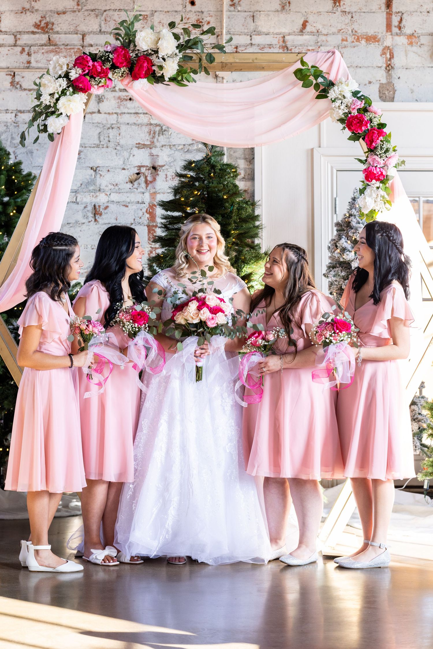 Wedding party gathered under a pink floral arch with bridesmaids in matching pink dresses.