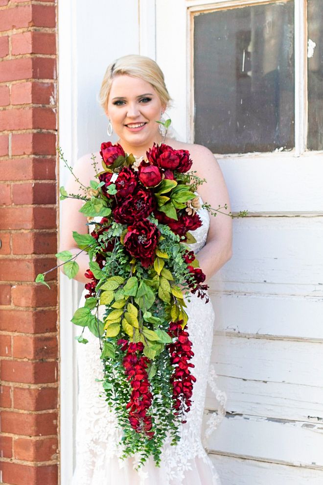 Bridal portrait with cascading burgundy and green bouquet against white door and brick wall.