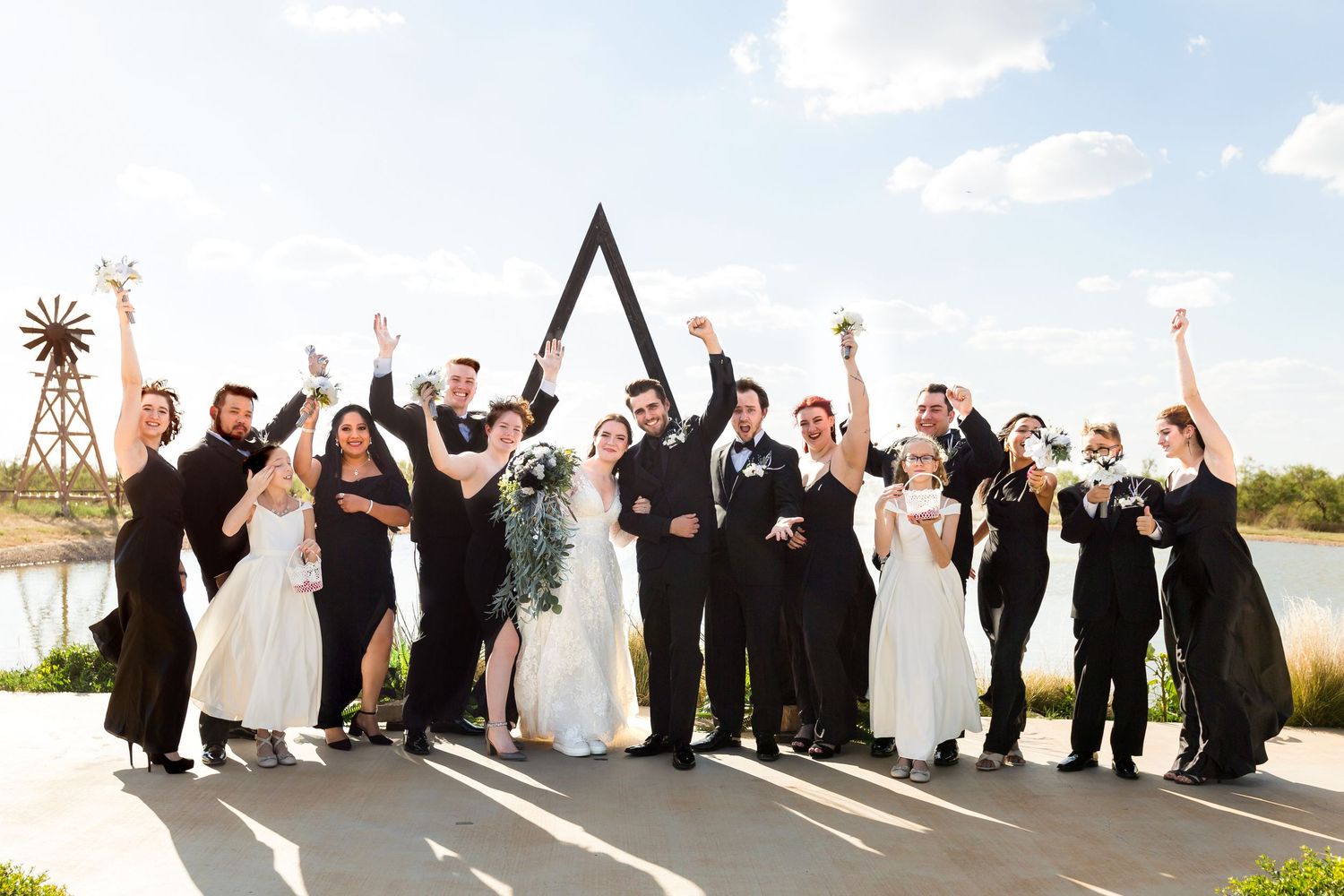 Wedding party celebrating with raised arms under a geometric arch structure outdoors.