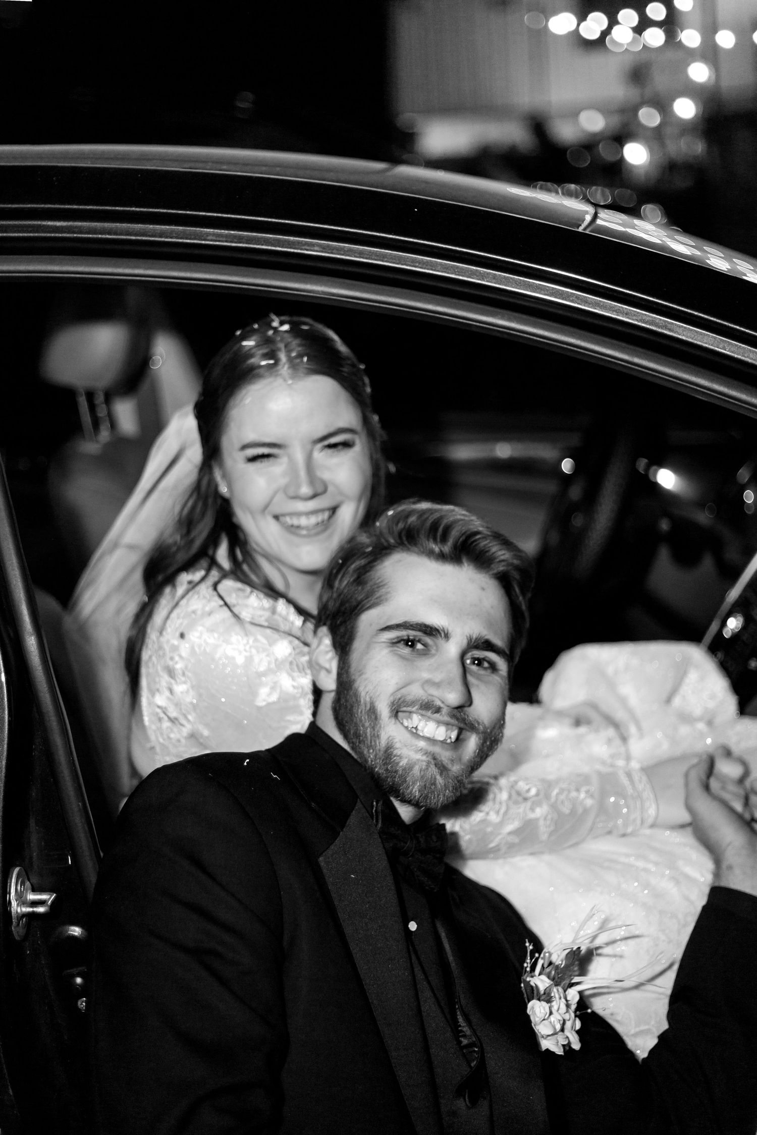 Newlyweds departing in car after wedding celebration with festive lights in background.