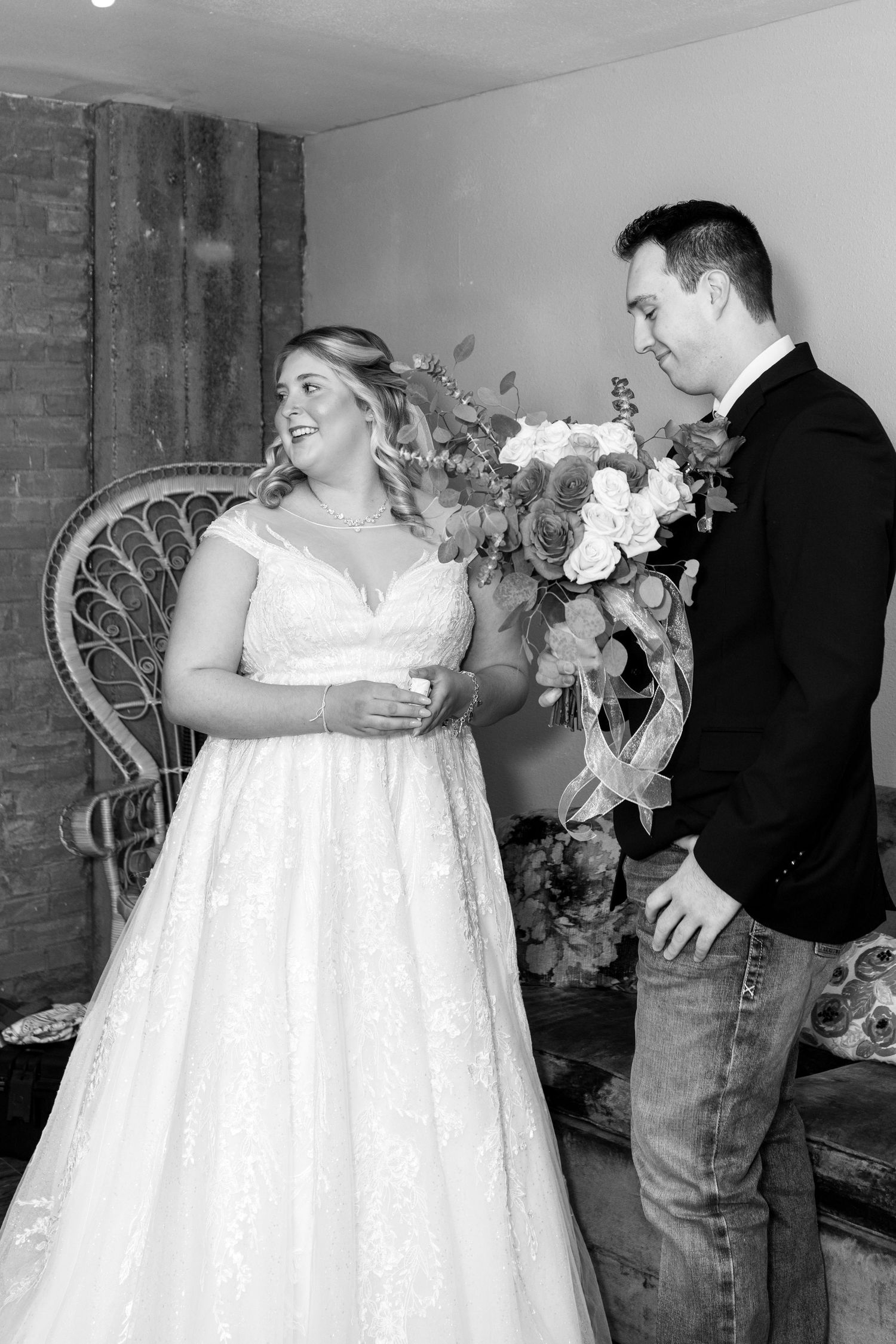 Black and white wedding photo with couple sharing a candid moment by vintage wicker chair.