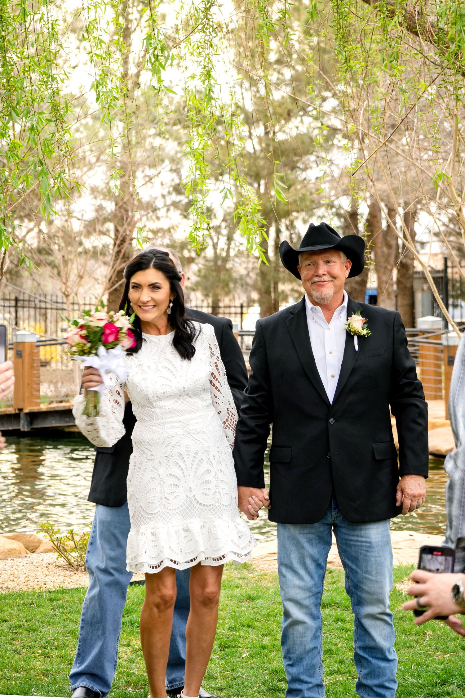 Wedding party posing on grass with willow trees and water in the background.
