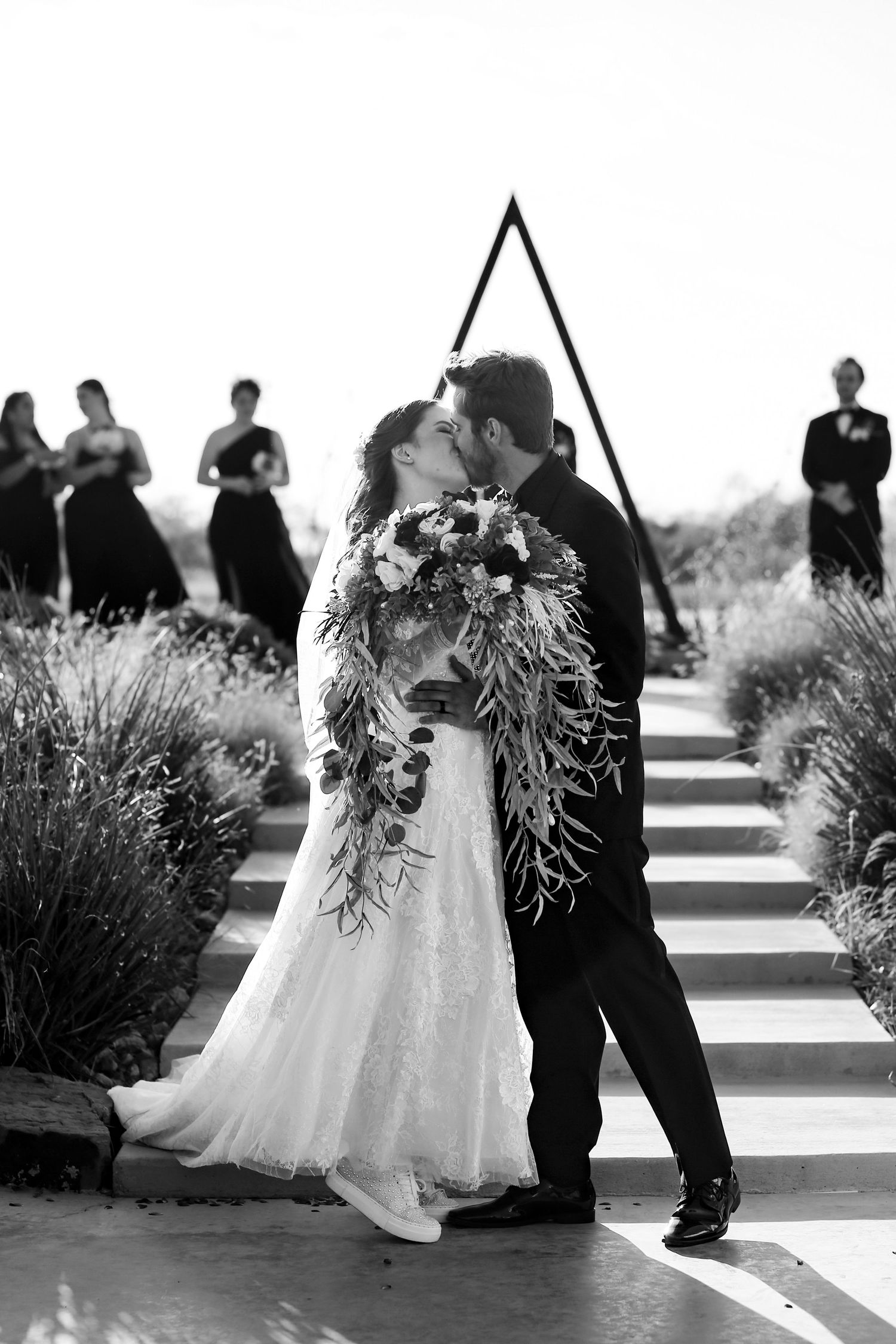Black and white ceremony photo with couple embracing under a triangular arch with bridal party in background.