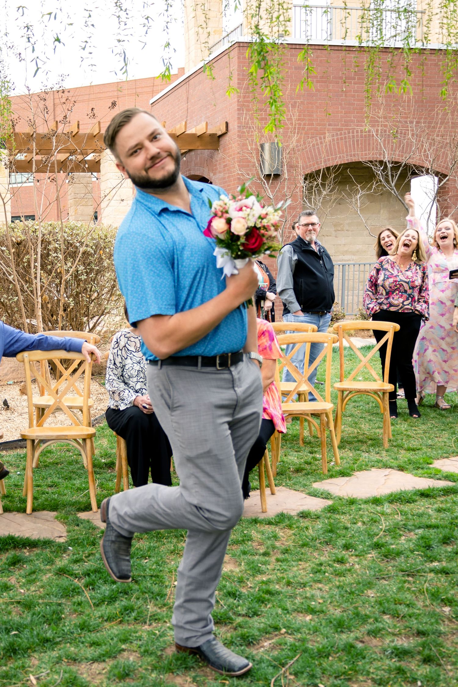 Playful groomsman performs silly dance moves on the grass at an outdoor wedding reception.