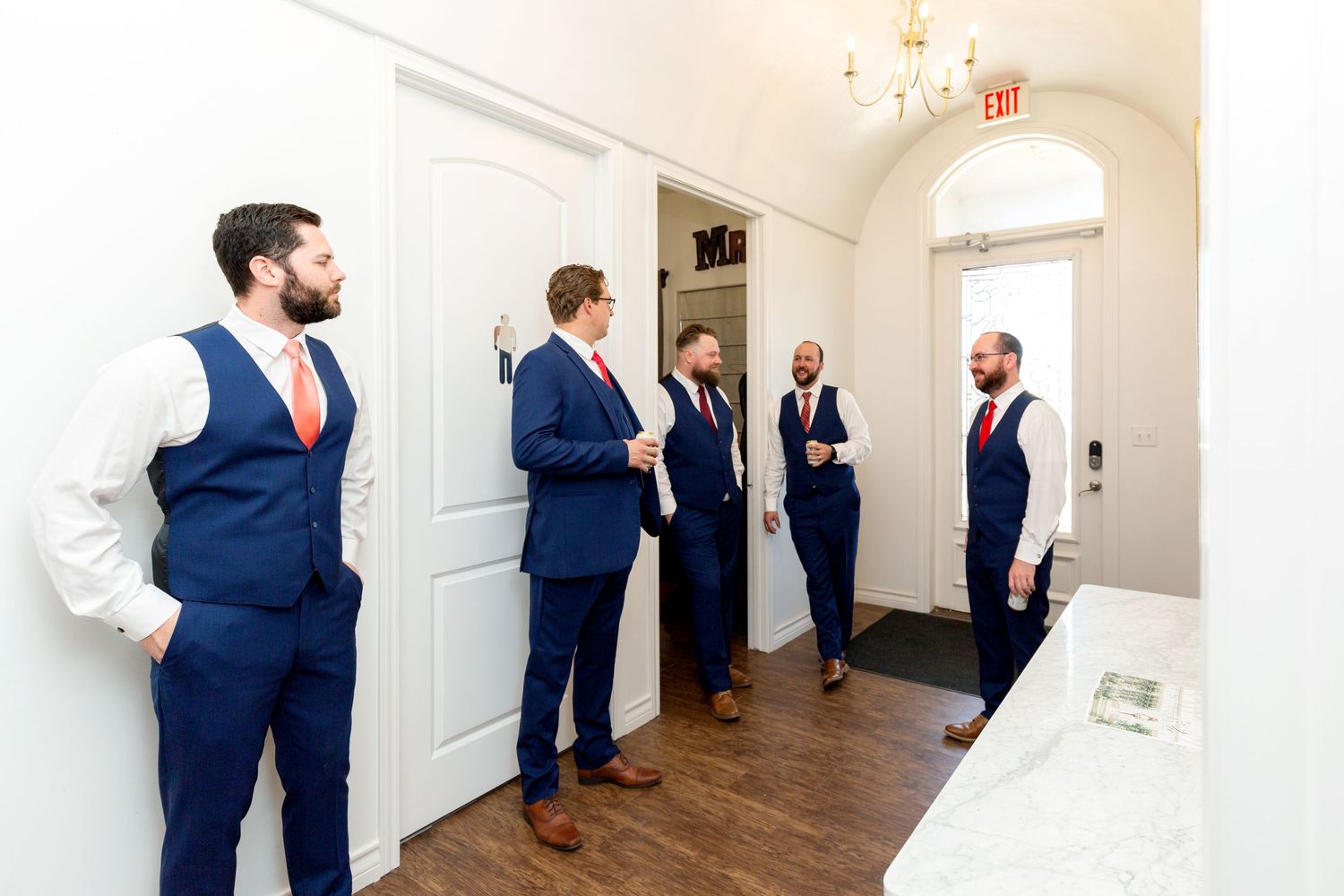 Row of groomsmen in navy suits standing in elegant white hallway before ceremony.