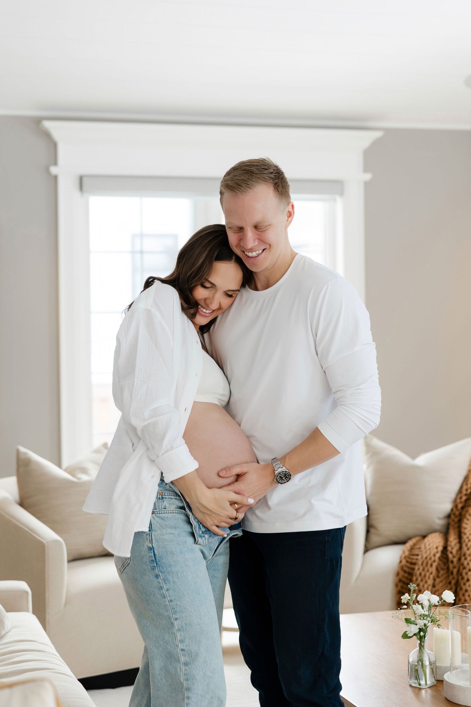 Couple shares intimate moment in bright white living room during maternity photoshoot.