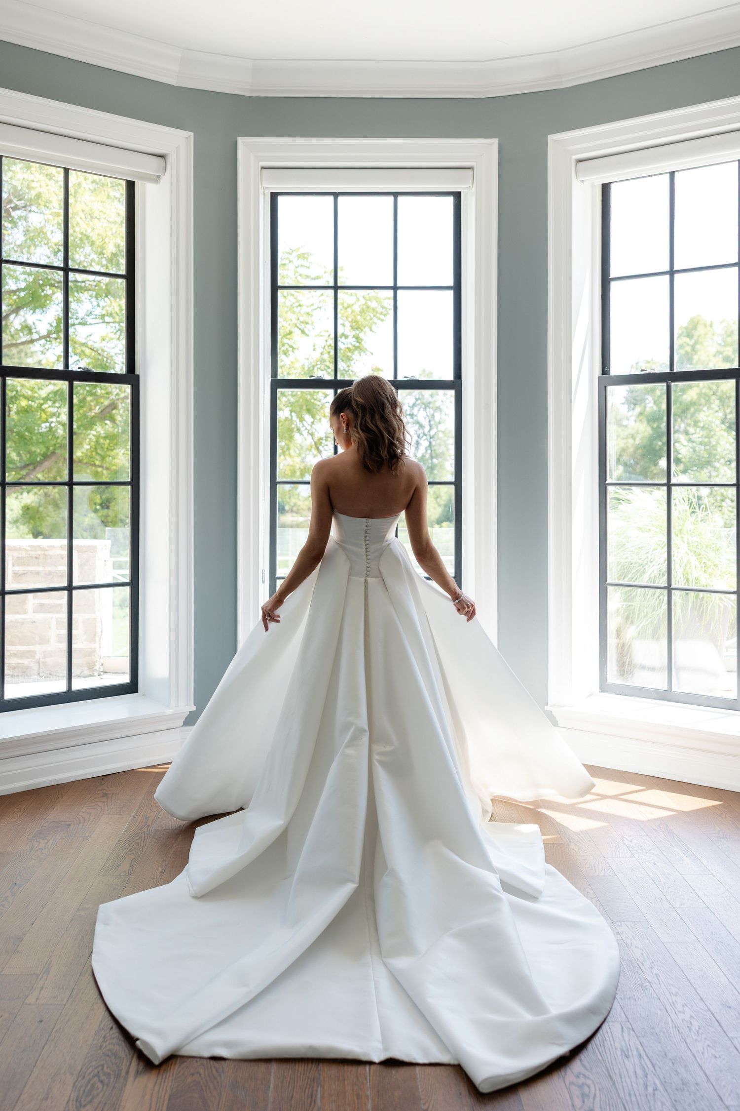 Elegant white wedding gown with dramatic train shown from behind in a bright corner room with large windows.
