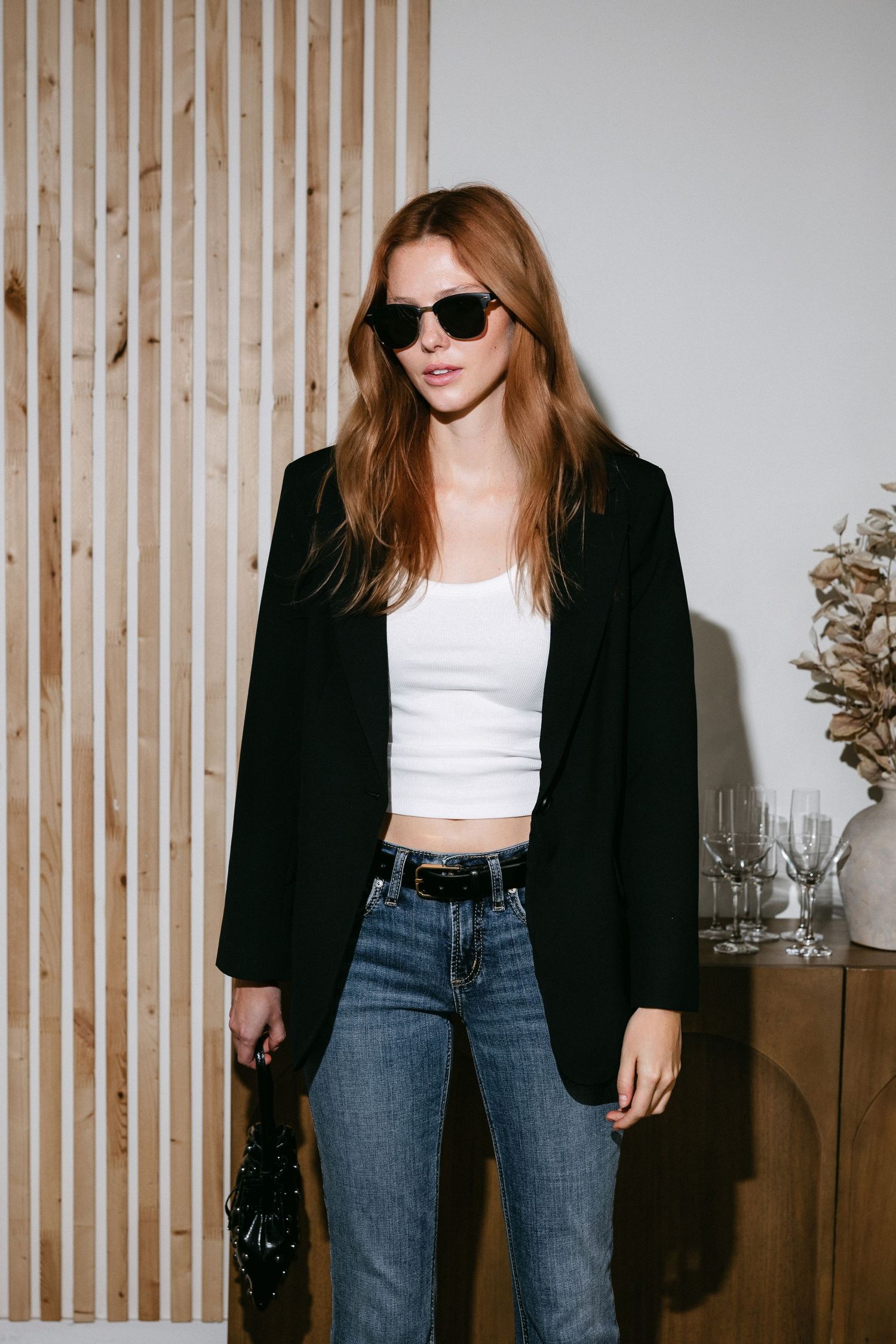 Fashion model wearing black blazer, white tank top and jeans against wooden slat wall background.