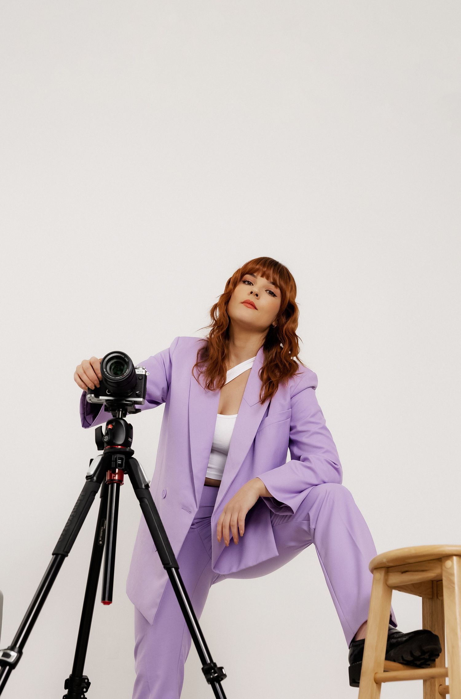 Fashion photographer in lavender blazer poses with professional camera equipment against white background.