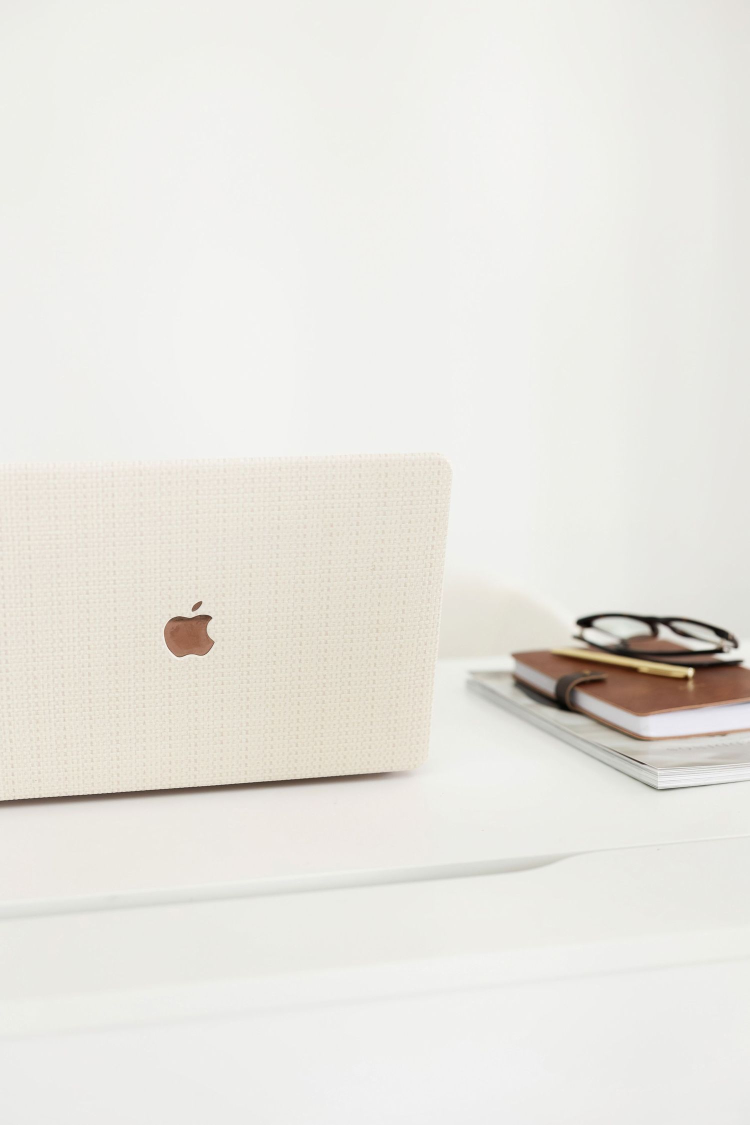 Minimalist workspace setup with MacBook laptop, glasses and notebooks on white desk.