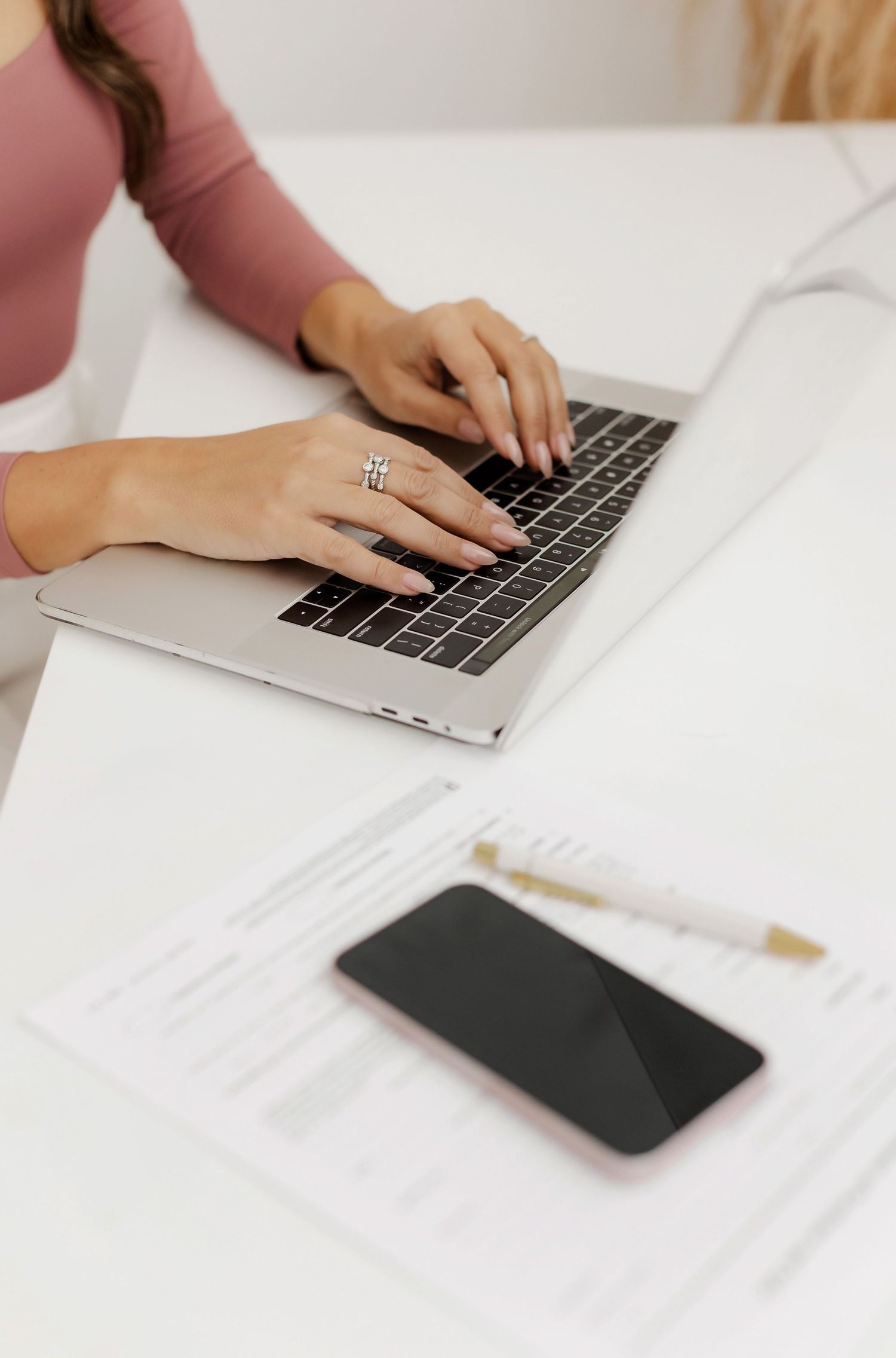 Close-up of hands typing on laptop keyboard on white desk with papers.