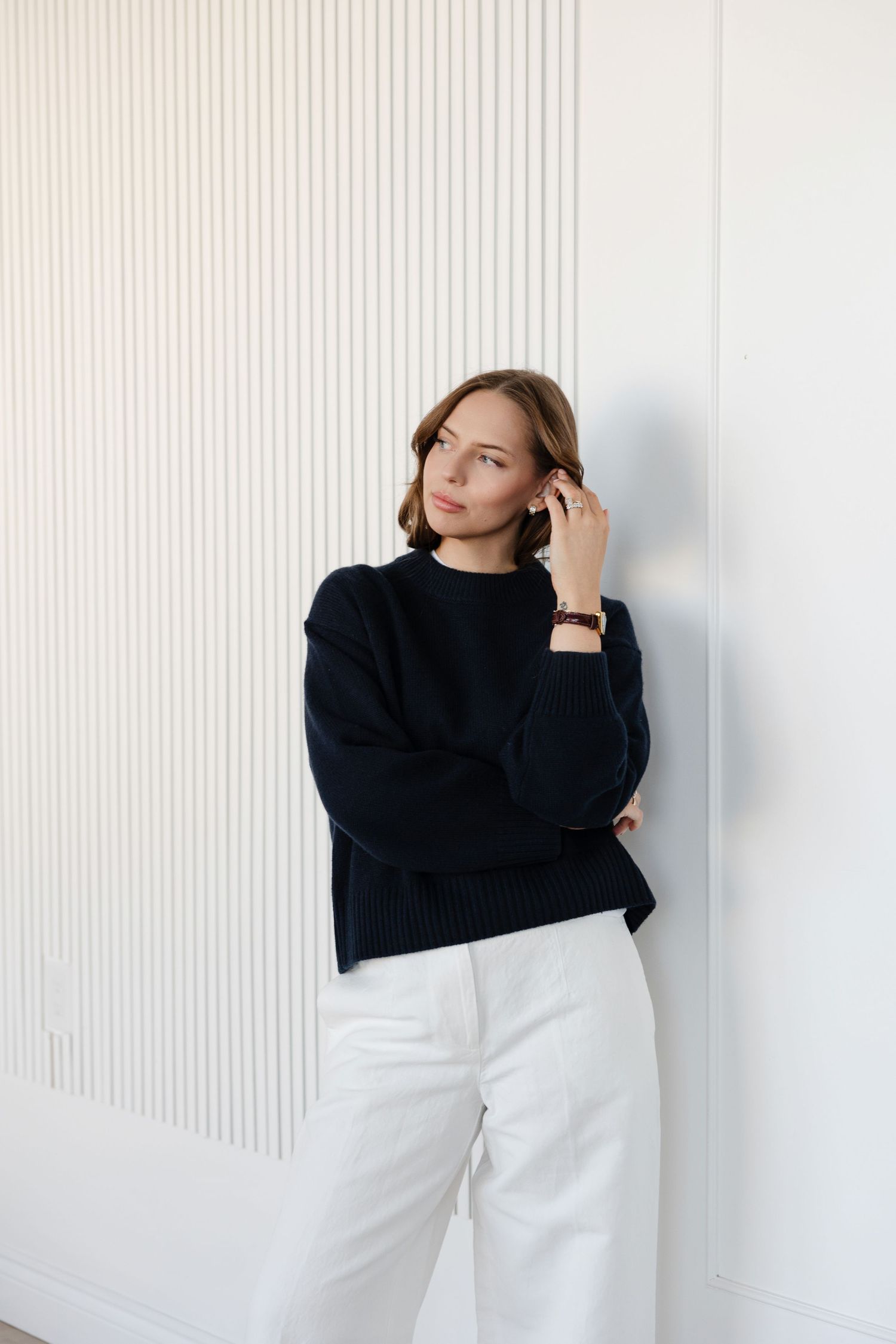 Model in black sweater and white pants poses against textured white wall in minimalist setting.