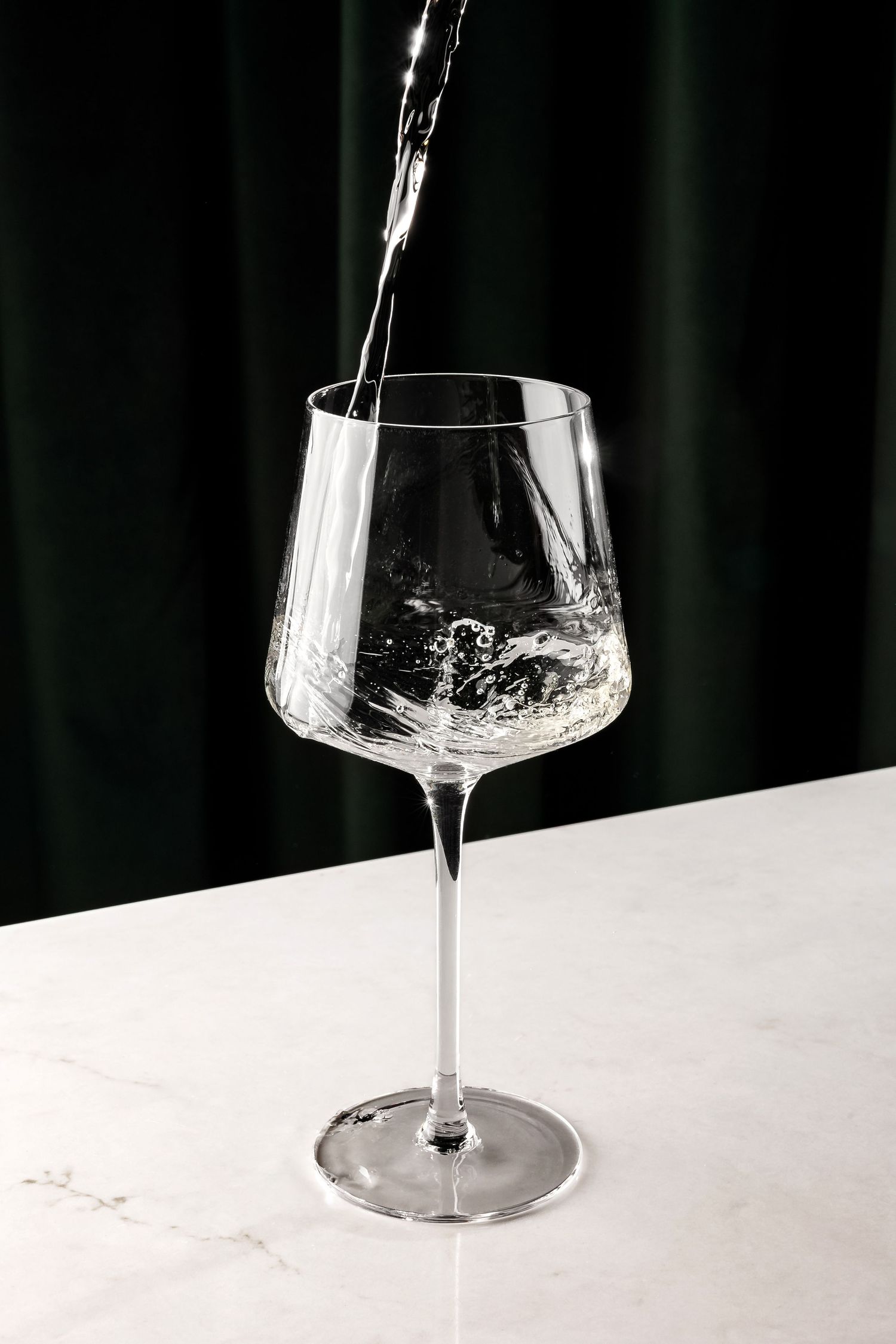 Crystal clear liquid streaming into an empty wine glass on white surface.