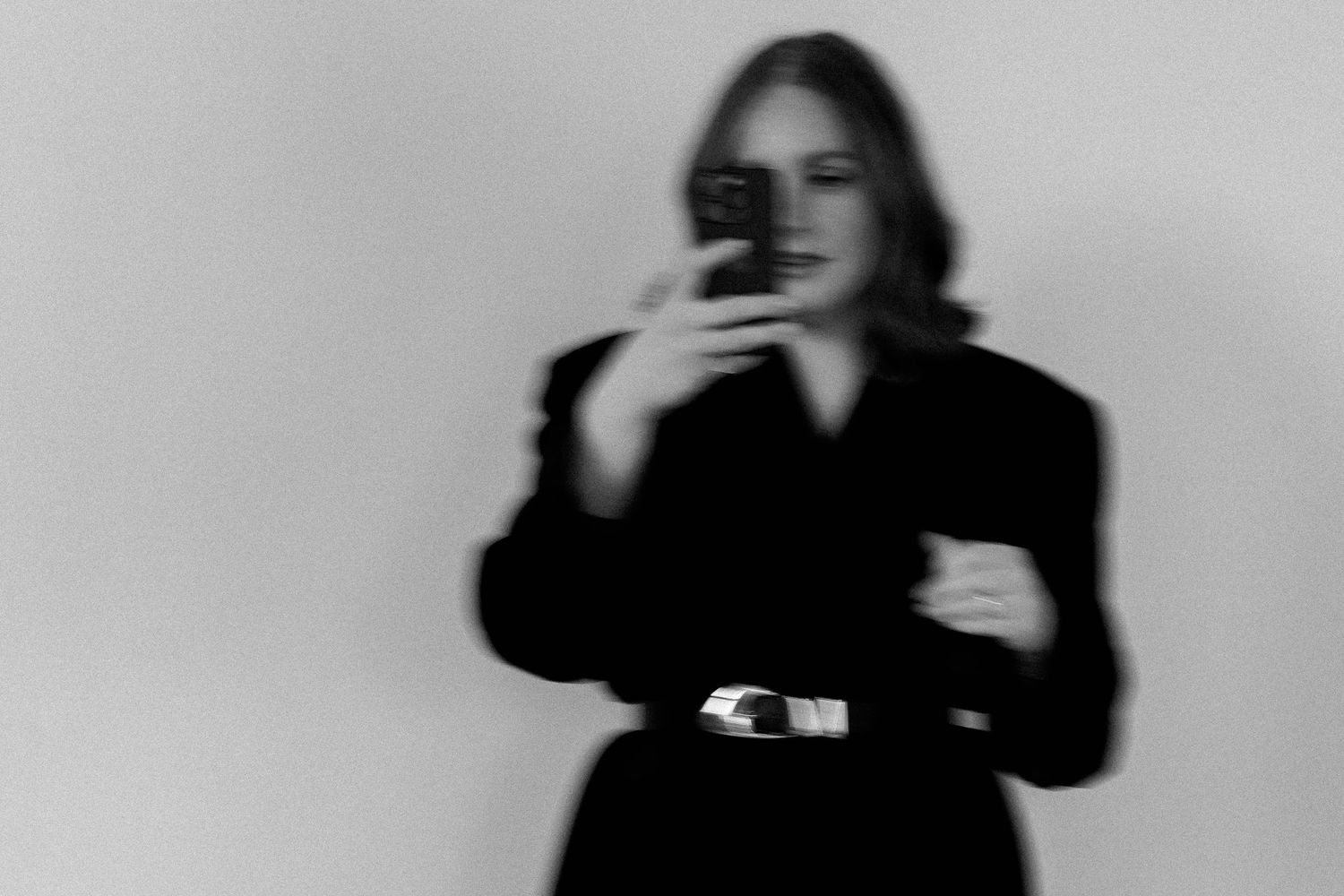 Person in dark clothing holds phone while posing dramatically against light background.