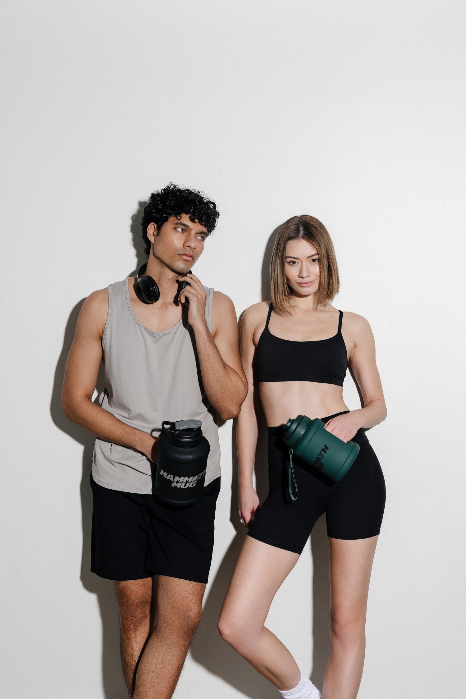 Two people in athletic wear pose against white wall with fitness accessories.