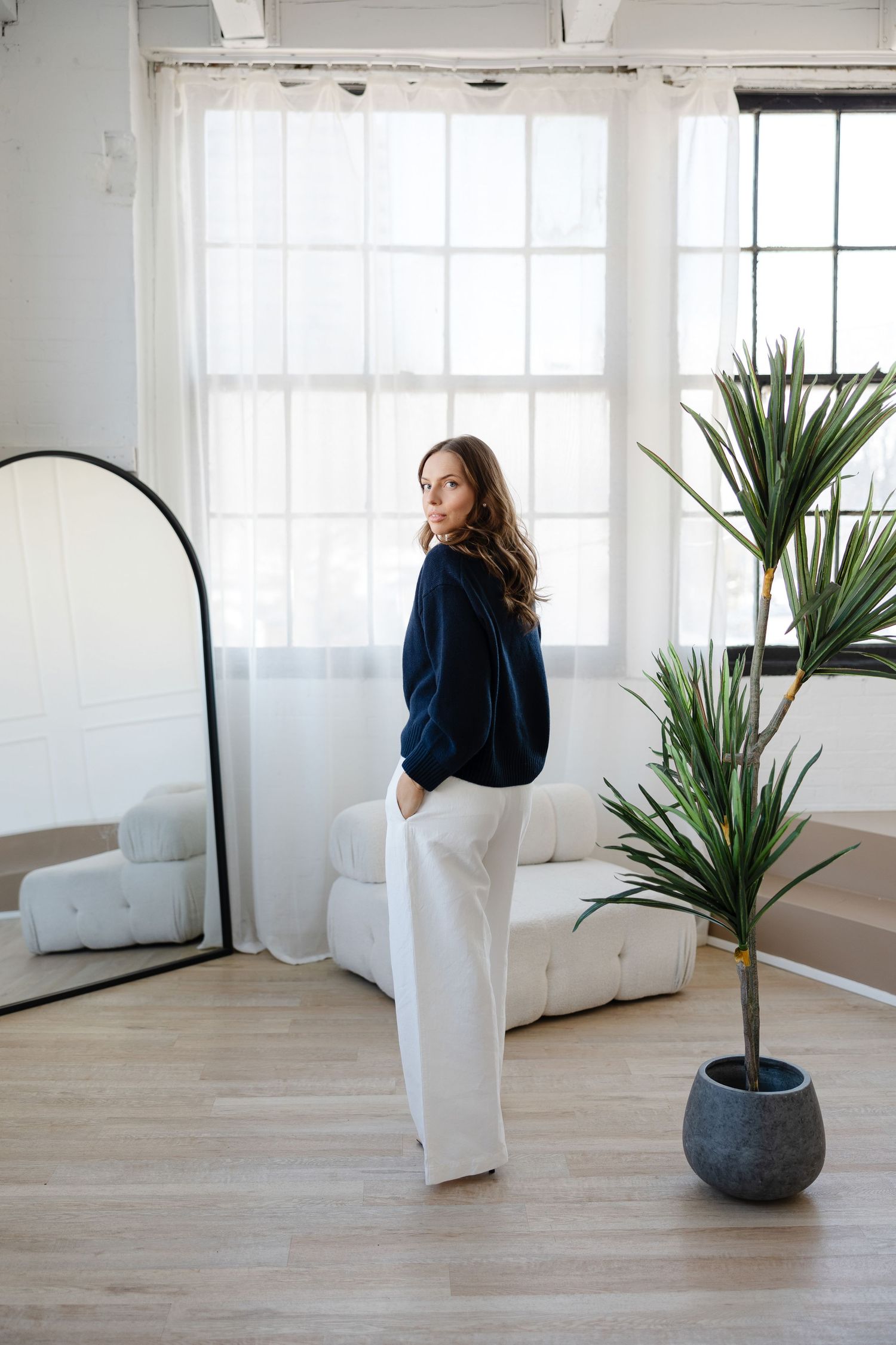 Bright modern room with large windows, potted plant, and figure in dark sweater and white pants.