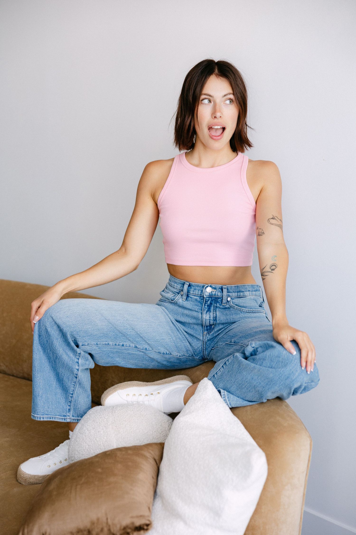 Relaxed pose in pink crop top and wide-leg jeans on beige couch.