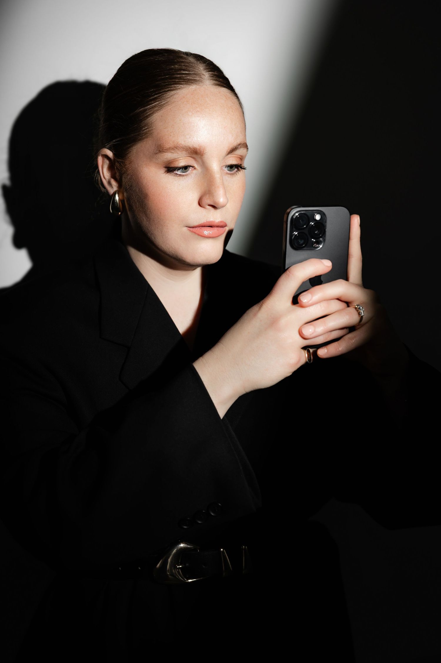 Dramatic portrait of someone in black blazer taking selfie with iPhone in moody lighting.