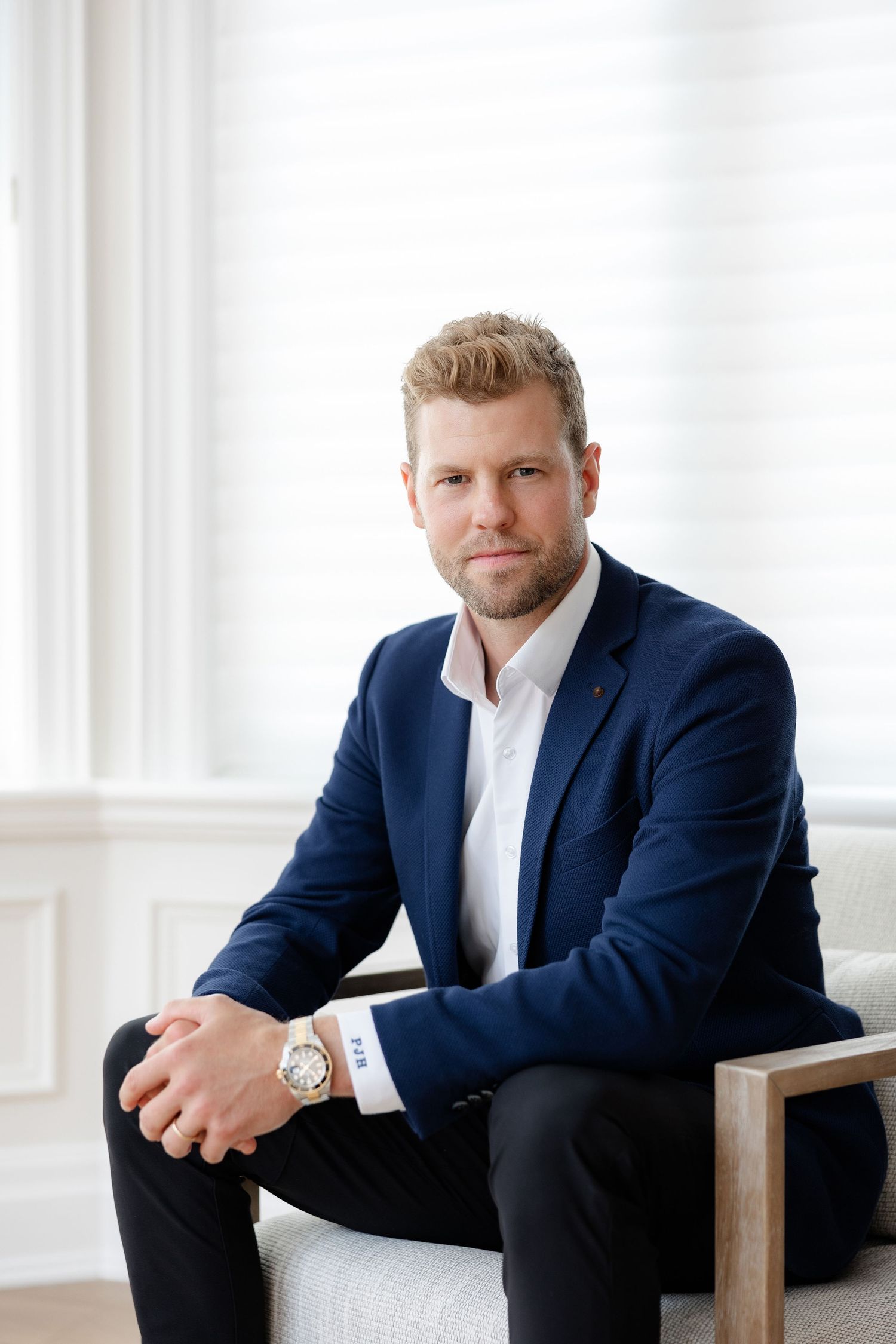 Professional business portrait of person in navy blazer and white shirt sitting in modern bright setting.