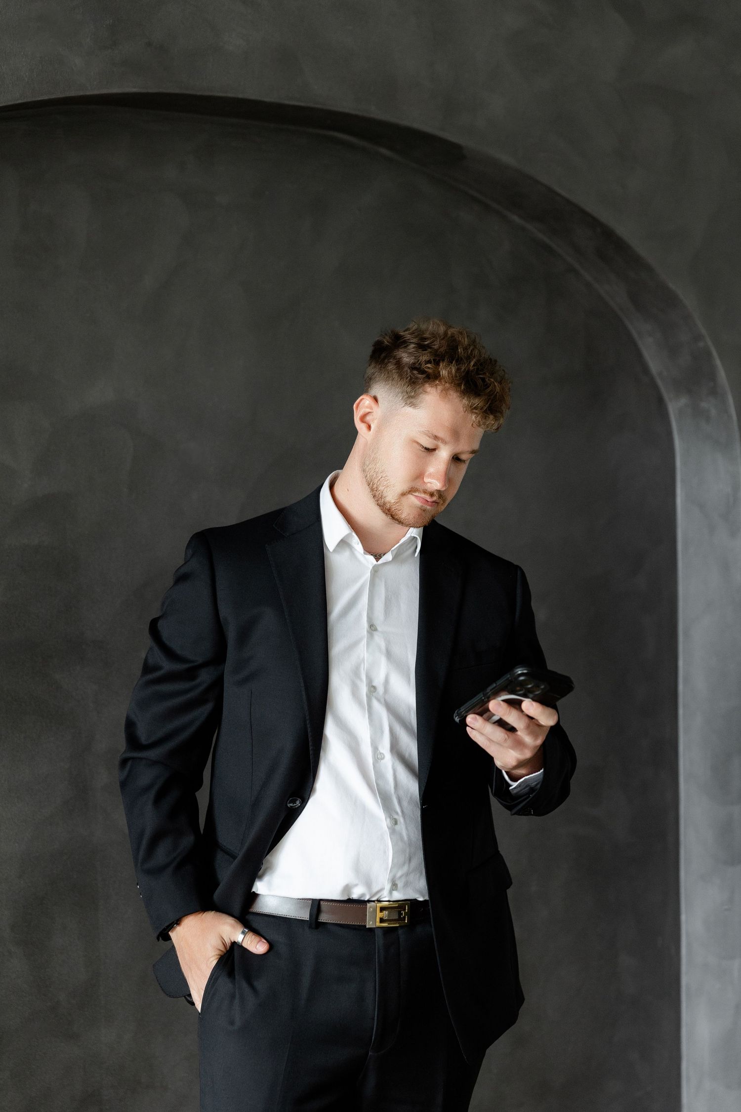 Professional in dark suit checks mobile phone against grey textured wall.