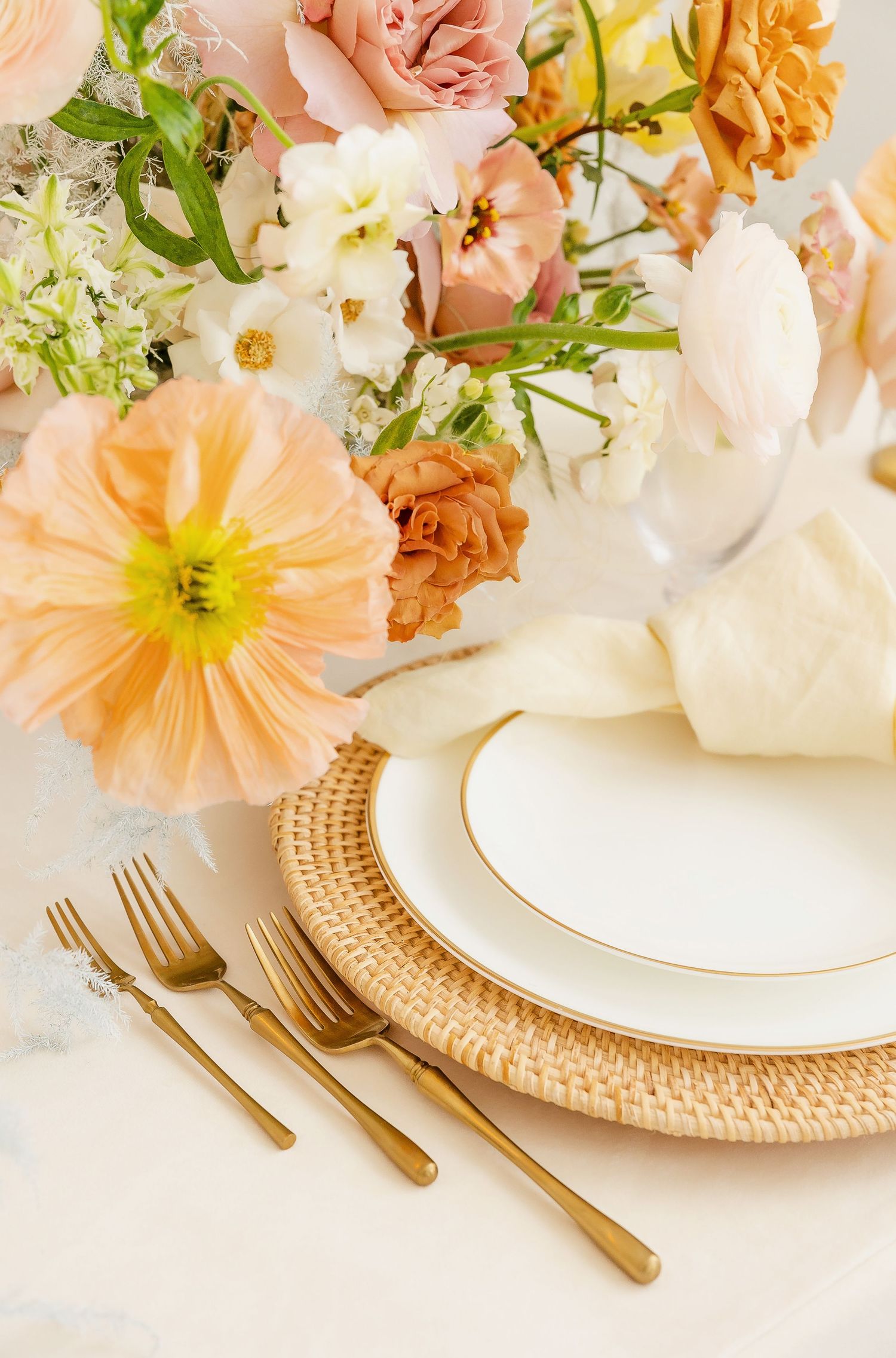 Formal table setting with gold-rimmed plates, woven chargers and peach floral arrangements.