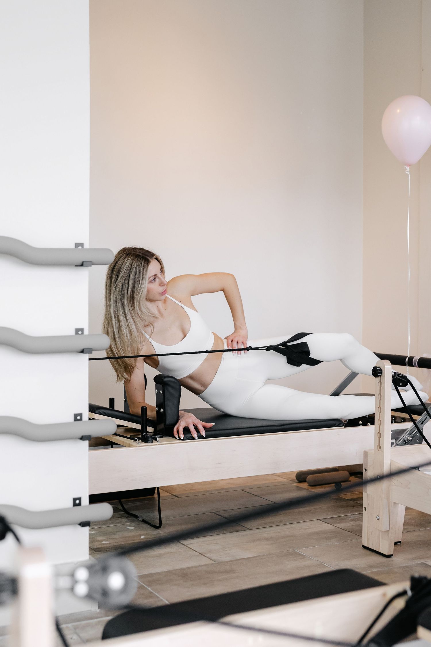 Person exercising on a Pilates reformer machine in a bright studio setting wearing white athletic wear.