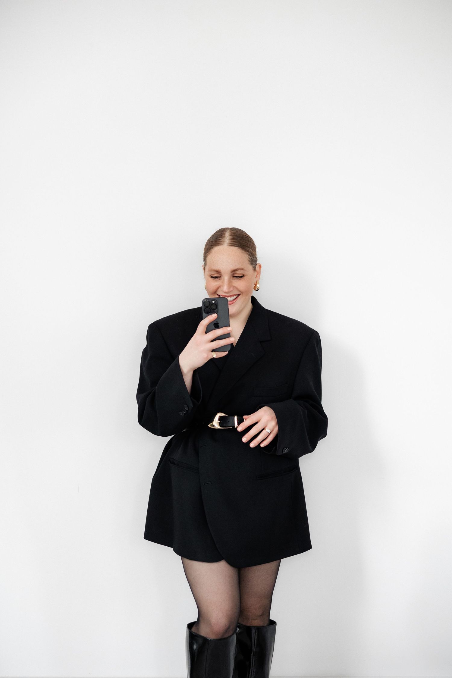 Stylish monochrome portrait of person in oversized black blazer and boots against white wall.