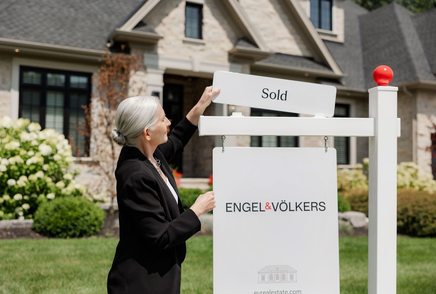 Real estate agent points to Engel & Volkers sold sign in front of stone residential property.