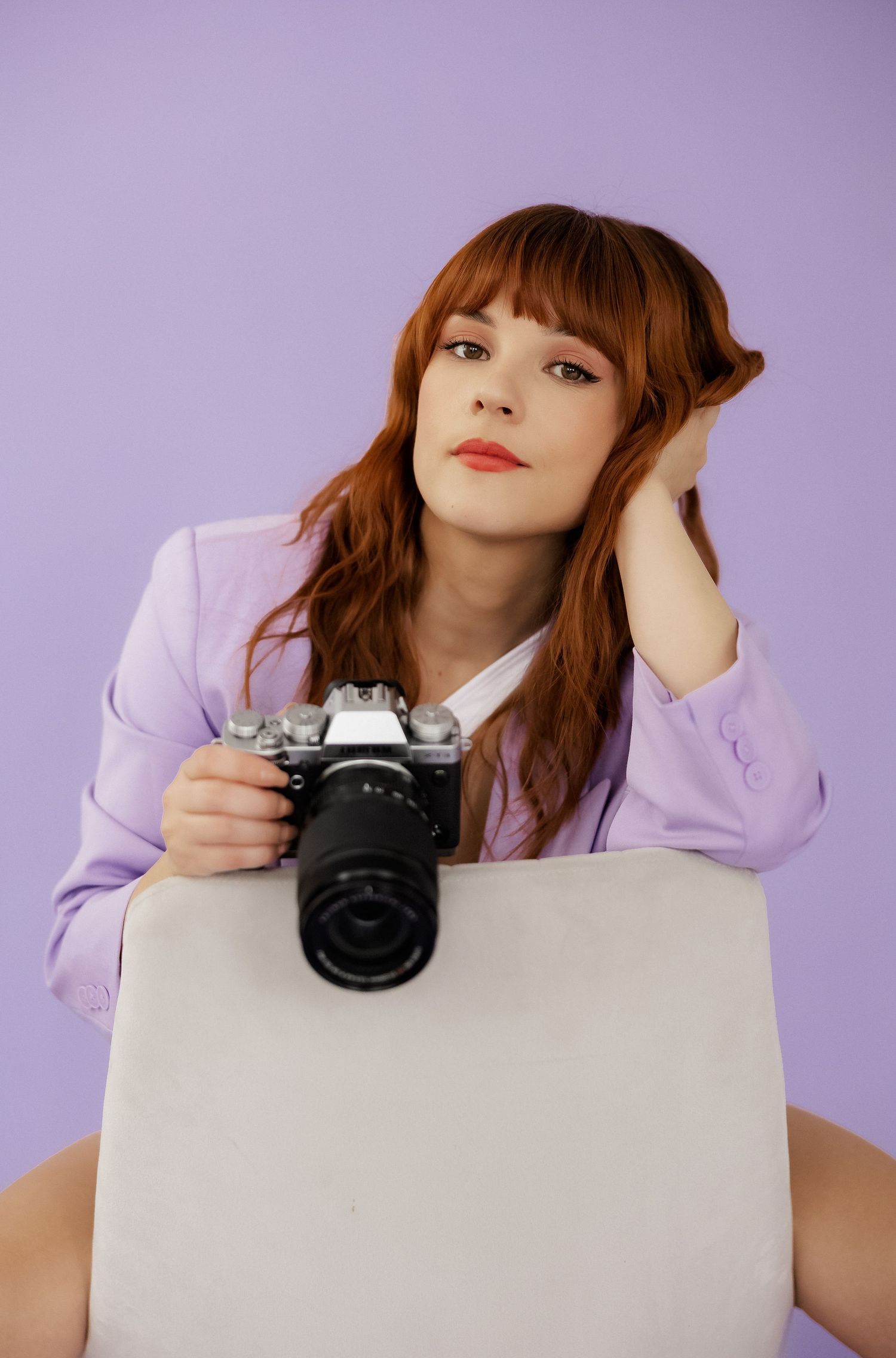 Fashion model with vintage camera poses on white cube in purple studio setting.